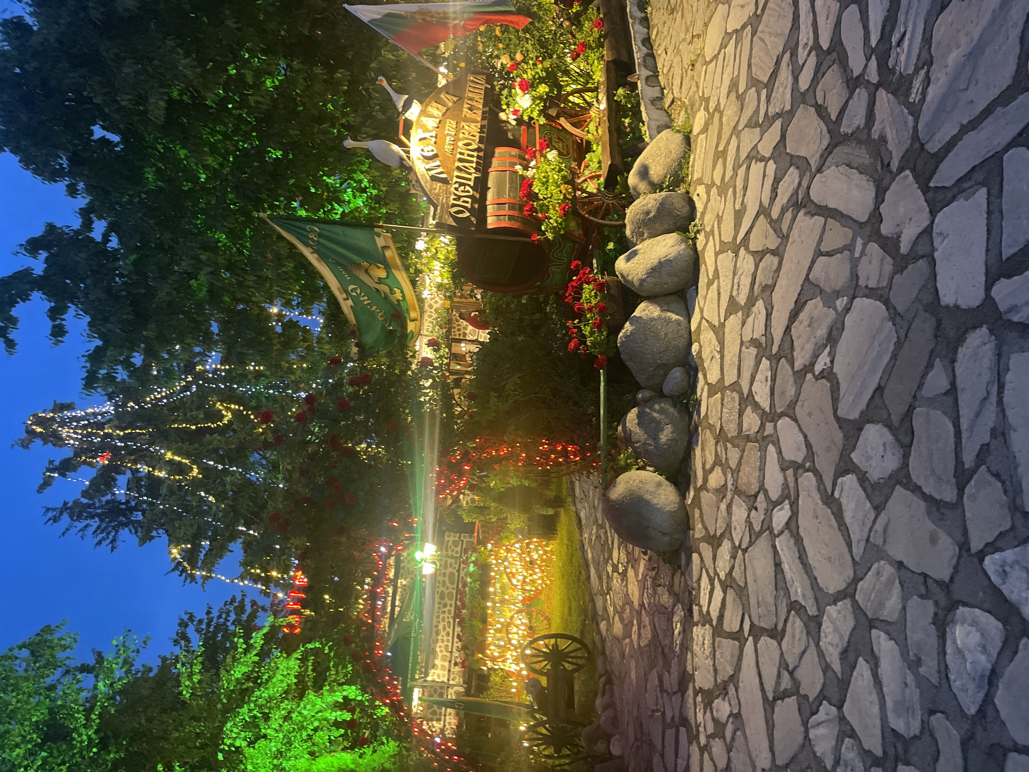 The image depicts an inviting outdoor scene at dusk or night, likely an entrance or garden area of a restaurant or tavern, illuminated by numerous artificial lights. A textured stone path or wall occupies the right side of the frame, bordered by a series of large, smooth rocks. The left side is dominated by rich green foliage, including trees and bushes, adorned with string lights glowing in white, yellow, and red hues, creating a warm and festive ambiance.
A central decorative feature is a rustic wooden sign, shaped like a barrel lid and framed by vibrant red and white flowers. Two white, stylized bird figurines, resembling cranes or storks, are perched on top of the sign. Below it, a wooden barrel is integrated into the landscaping, also surrounded by red blooms. The sign prominently displays Cyrillic text: "МЕХАНА" (Mehana, meaning "Tavern"), "ОБЩАНОВА КЪЩА" (Obshtanova Kushta, meaning "Obshtanova's House"), and "ОТ 1772" (Since 1772), indicating an old, traditional establishment.
Further to the left, a green flag with an emblem and partially visible Cyrillic text (e.g., "СВО" and "ew.pb.BG") hangs, along with one or more Bulgarian national flags (white, green, red stripes), confirming the location as Bulgaria. A decorative wooden wagon wheel is visible on the ground amidst the glowing lights and foliage. The dark blue sky confirms the evening hour. The scene, devoid of people, projects a welcoming, traditional, and cozy atmosphere, characteristic of a Bulgarian "mehana."