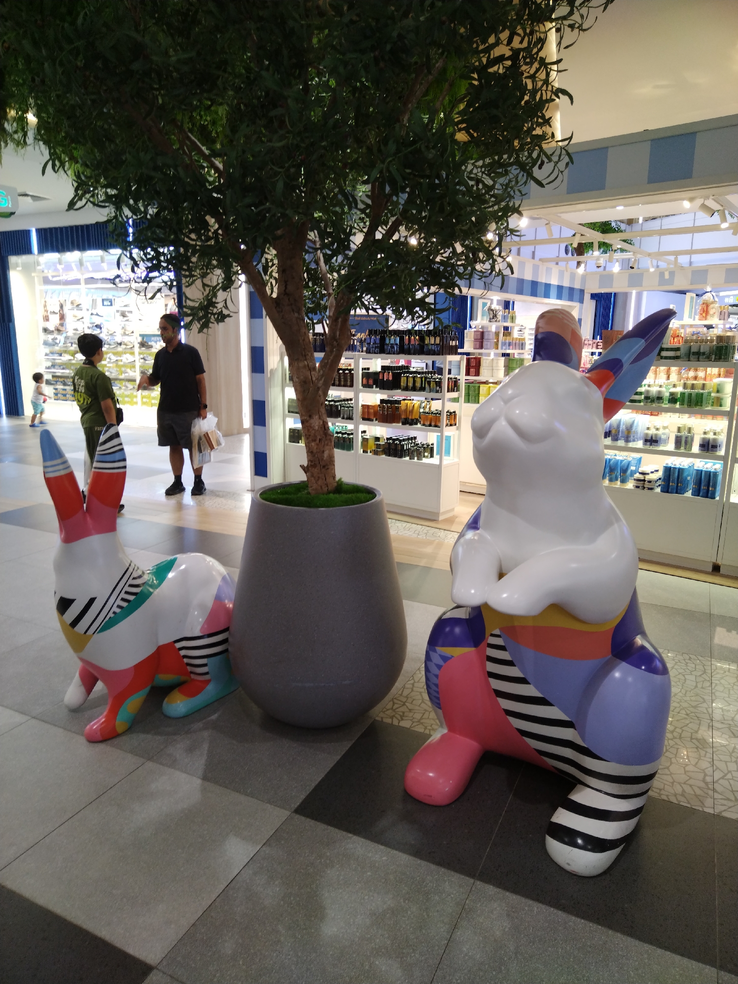 The image shows two colorful rabbit sculptures in front of a store in Patong, Thailand. The sculpture on the left is white with red, green, blue, yellow, and black geometric patterns. The sculpture on the right is also white with blue, purple, pink, and black striped patterns. Both sculptures are life-sized and have large ears.
In the background, there are shelves stocked with various products, likely in a retail store. Two men and a child are standing near the entrance to the store. One man, wearing a black polo shirt and shorts, is holding a plastic bag. The other man is wearing a green t-shirt and shorts. The child is wearing a blue and white striped shirt and blue shorts. A large potted plant with green foliage is situated between the sculptures. The floor is tiled with a checkered pattern of light and dark gray squares. The lighting suggests an indoor environment, possibly a mall or a large retail establishment.