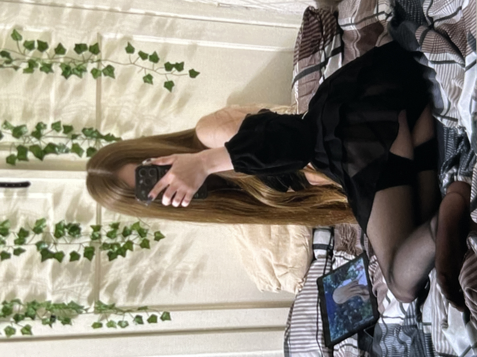 A person with long, auburn hair sits on a bed, holding a smartphone in front of their face, obscuring it. They are wearing a black, off-the-shoulder top with ruffled sleeves and a layered black skirt, as well as black stockings. To the left of the person, a tablet displays an image of an anime character. The bed is covered with a comforter and pillows. In the background, a white wall is decorated with artificial ivy vines. The scene appears to be set in a bedroom.