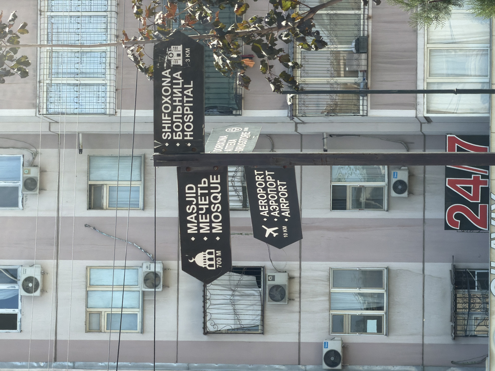 The image is a street view in Tashkent, Uzbekistan, showing a building facade with multiple windows and air conditioning units. A directional signpost stands in front of the building, with black signs pointing in different directions. The signs have text in Uzbek, Russian, and English. One sign indicates "MASJID / МЕЧЕТЬ / MOSQUE 700 M". Another sign indicates "AEROPORT / АЭРОПОРТ / AIRPORT 10 KM". A third sign indicates "SHIFOXONA / БОЛЬНИЦА / HOSPITAL 3 KM". A fourth, smaller sign points to "DORIXONA / АПТЕКА / DRUGSTORE". A tree with sparse leaves is visible to the right of the signpost, partially obscuring the building. Below the signs, part of a red and white sign with the numbers "24/7" is visible. The overall scene depicts an urban environment with clear signage for various destinations.