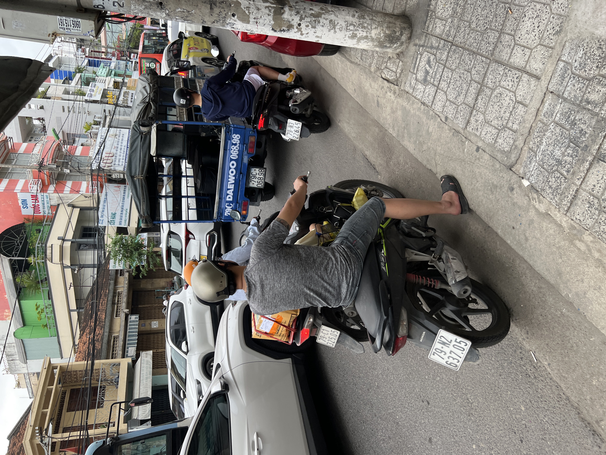 This is a medium-shot, eye-level, horizontal photograph of a busy street scene in Nha Trang, Vietnam, with a focus on traffic and urban infrastructure. The foreground features a man riding a black motorcycle, seen from the rear. He is wearing a grey t-shirt, denim shorts, and black flip-flops, with a gold helmet and an orange object, possibly another helmet, on the back of his motorcycle. A cardboard box is strapped to the rear of his motorcycle. The motorcycle's license plate reads "79-C1 325.05". To the left, the front of a white car is visible, and further back, a white SUV. Behind the motorcycle, a blue, open-bed truck is prominent, with a license plate "79C DAO EWO 068.98". A person in a navy blue hoodie and white shorts is riding on the back of this truck, wearing a black helmet. To the right, a yellow taxi is partially visible, and a red car is parked or stopped by the roadside. The street is paved with asphalt, and a concrete sidewalk with decorative tile patterns runs along the right side. The background is filled with multi-story buildings of various architectural styles and colors, typical of Vietnamese urban environments. Electrical wires are strung across the street, and various signs are visible on the buildings, some with Vietnamese text. The sky is overcast. The overall scene depicts a typical moment of urban traffic congestion in Vietnam.