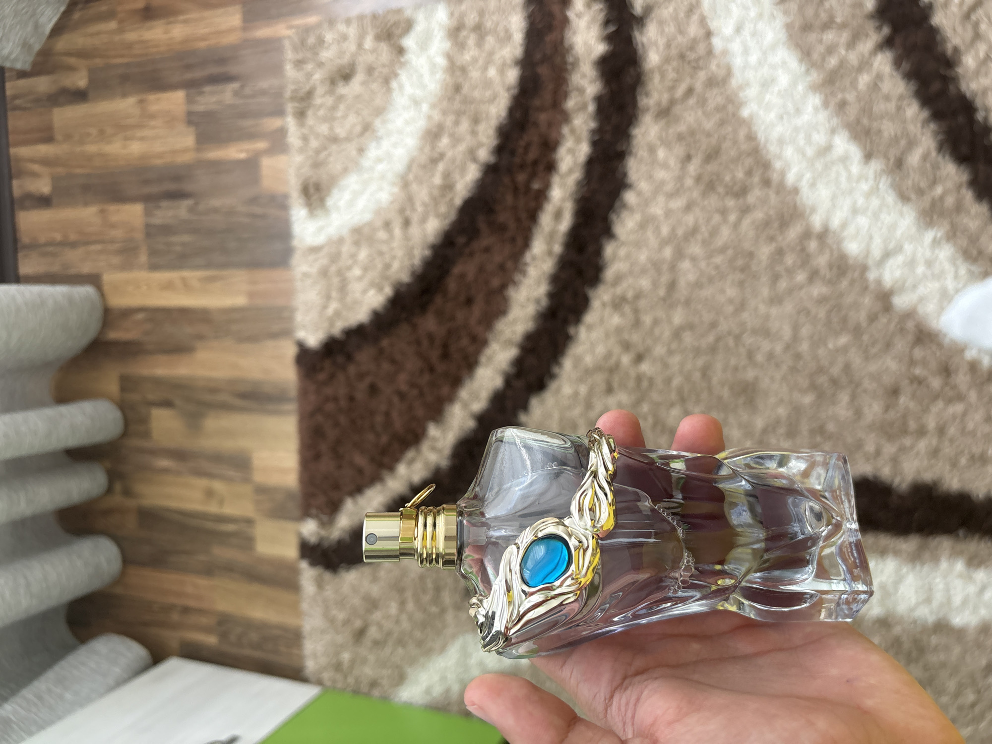 A hand holds a glass perfume bottle with a gold sprayer and a decorative gold-colored embellishment featuring a blue stone. The background features a brown and beige striped rug and wood-look flooring. The location in the city of Stara Zagora, Bulgaria cannot be determined from the image.
