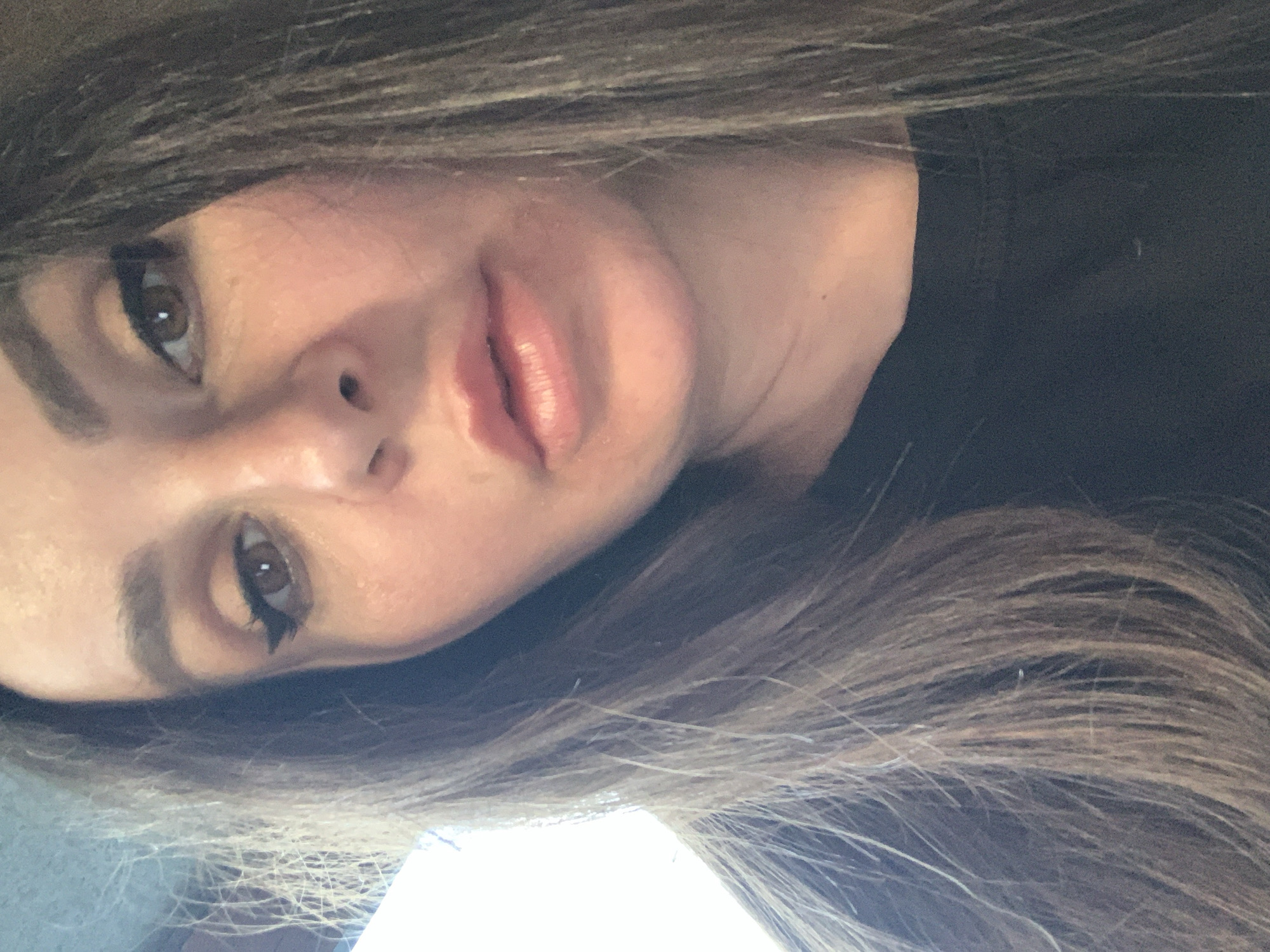 A close-up, rotated selfie features a person, likely a woman, with her head tilted to the side, looking directly at the camera. Her long, multi-toned brown hair with lighter highlights frames her face and flows downwards towards the right of the frame. She has medium brown eyes accentuated with dark, winged eyeliner and well-defined eyebrows. Her lips are full and appear to have a light pink or nude lip color. The skin has a natural texture. A dark garment, possibly black, is visible on her upper right side. The background is largely dark and out of focus, with a brighter, out-of-focus area towards the bottom left. The lighting is soft, and her expression is neutral. There is no visible text in the image.
