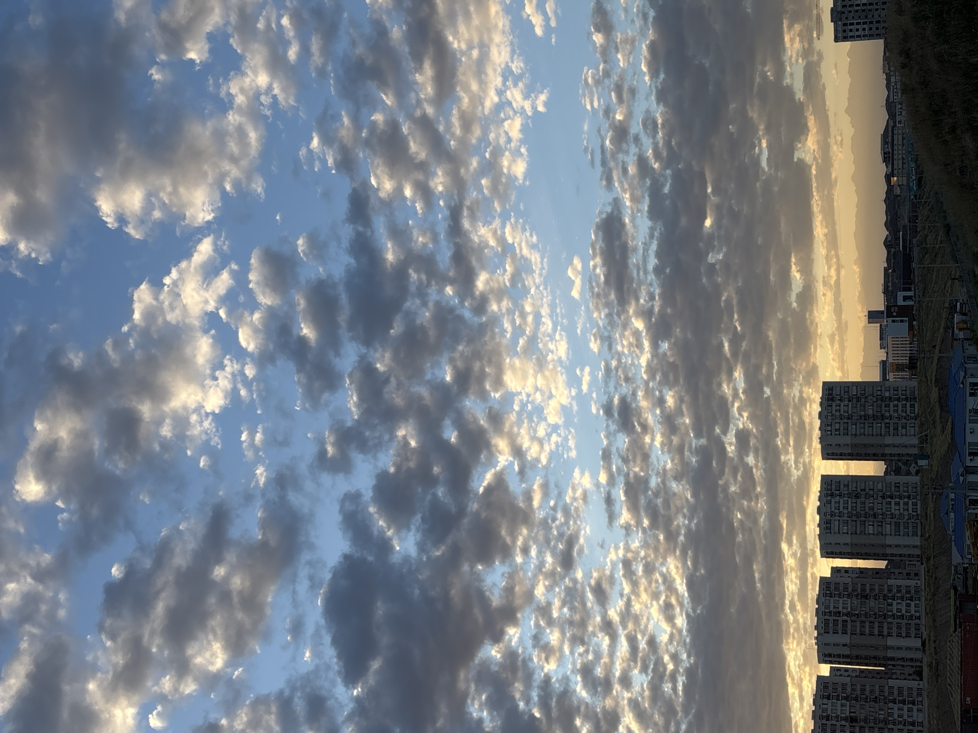 The image depicts a skyline view from a distance, likely from an elevated position on a hill, overlooking a city. The sky is filled with scattered clouds, with patches of blue sky visible. The clouds are illuminated by a warm, golden light, suggesting either sunrise or sunset. In the foreground, there are some sparsely vegetated hills and what appear to be construction sites with some materials and structures.
Dominating the middle ground are several tall, apartment buildings, all of similar design, featuring multiple stories with many windows. These buildings are clustered together. Further in the background, more of the city's urban landscape is visible, with a mix of smaller buildings and structures. The overall impression is of a developing urban area with a dramatic sky. The image does not show any individuals or specific actions. The scene appears to be located in Shymkent, Kazakhstan, judging by the architectural style of the apartment buildings and the natural landscape.