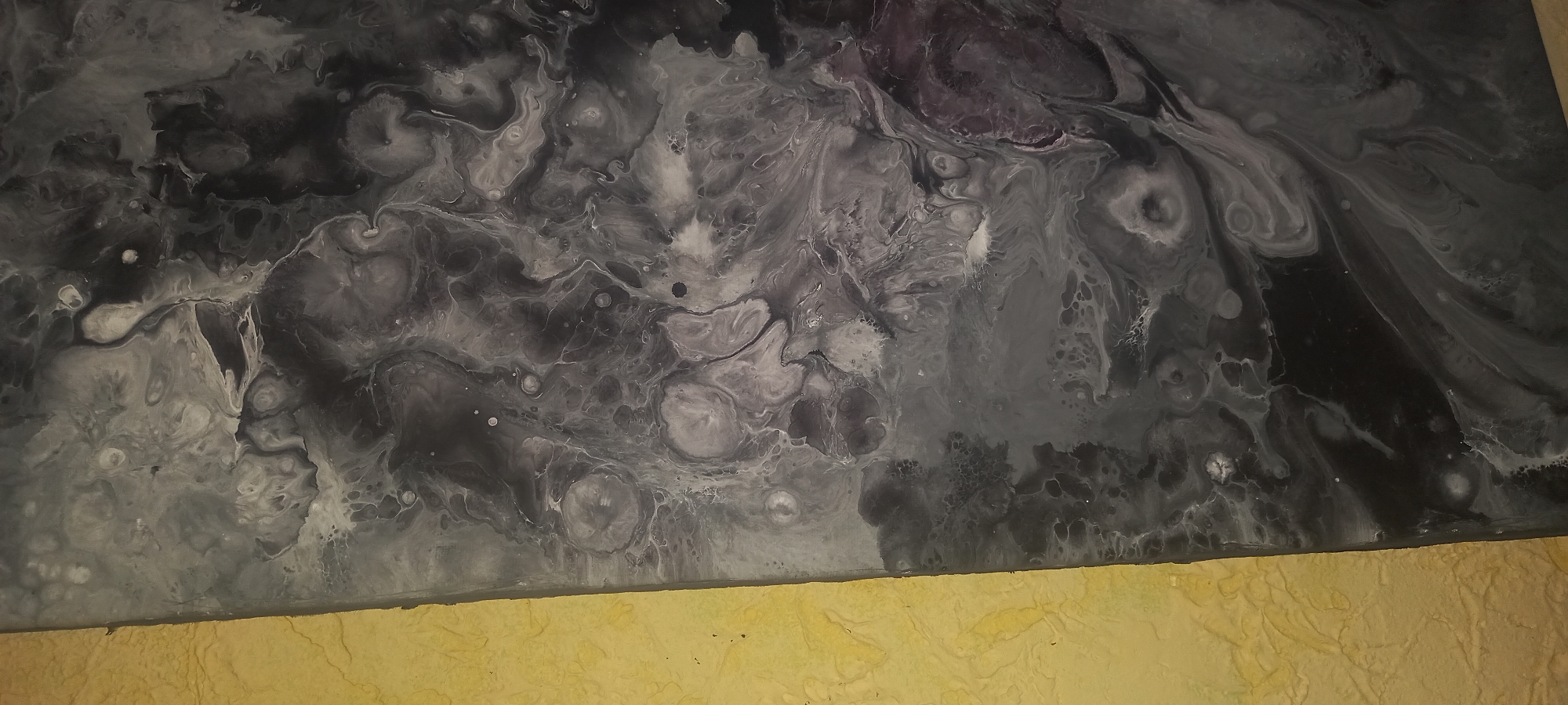 The image depicts an abstract painting with a swirling mix of gray, white, and black shades. The upper portion of the image is dominated by the painting, which has a marbled appearance with irregular patterns and textures. Below the painting is a section of a textured, yellow-colored surface, possibly a wall or table. The scene appears to be indoors, but there are no discernible human subjects or specific objects to pinpoint the location in Kryvyi Rih, Ukraine.