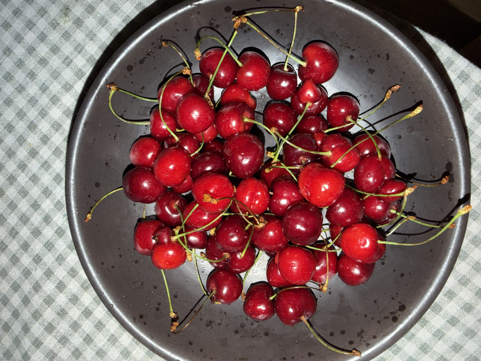 A high-angle, close-up shot depicts a dark gray bowl filled with a pile of red cherries with green stems. The bowl is positioned on a surface covered with a light gray and white checkered tablecloth. The scene appears to be indoors, illuminated by artificial lighting. The image does not include any people, specific landmarks, or contextual information that would identify the location as Khimki, Russia.