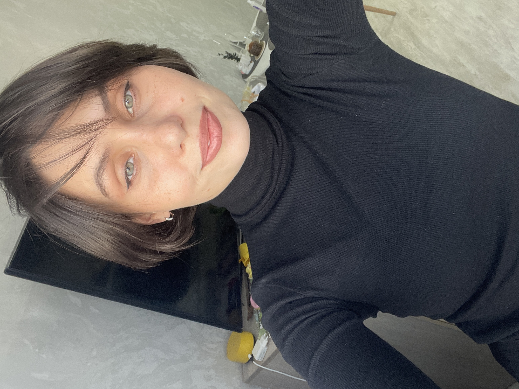 A portrait-oriented photograph, rotated approximately 90 degrees counter-clockwise, captures a young adult woman with short, dark brown hair and light green eyes. She is positioned in the foreground, looking directly at the camera with a gentle, faint smile. She is wearing a black, ribbed turtleneck top with long sleeves. Her eyes are accented with dark eyeliner, and her lips have a soft pink or nude shade. Small silver hoop earrings are visible in her ears.
The background indicates an indoor setting, likely a casual room within a residence in Izhevsk, Russia. To the left of the woman, a dark, rectangular object resembling a turned-off television or monitor is partially visible. Below and behind this object, a light grey, subtly textured wall forms part of the background. To the right of the screen, the area appears somewhat cluttered, featuring a bright yellow sponge, white cables, and various other indistinct items resting on a surface or shelf. Further back, small plants or decorative objects in containers are discernible, along with a light grey, textured surface, possibly a floor or tabletop. The lighting is soft and even, suggesting indoor ambient light. There is no visible text in the image.