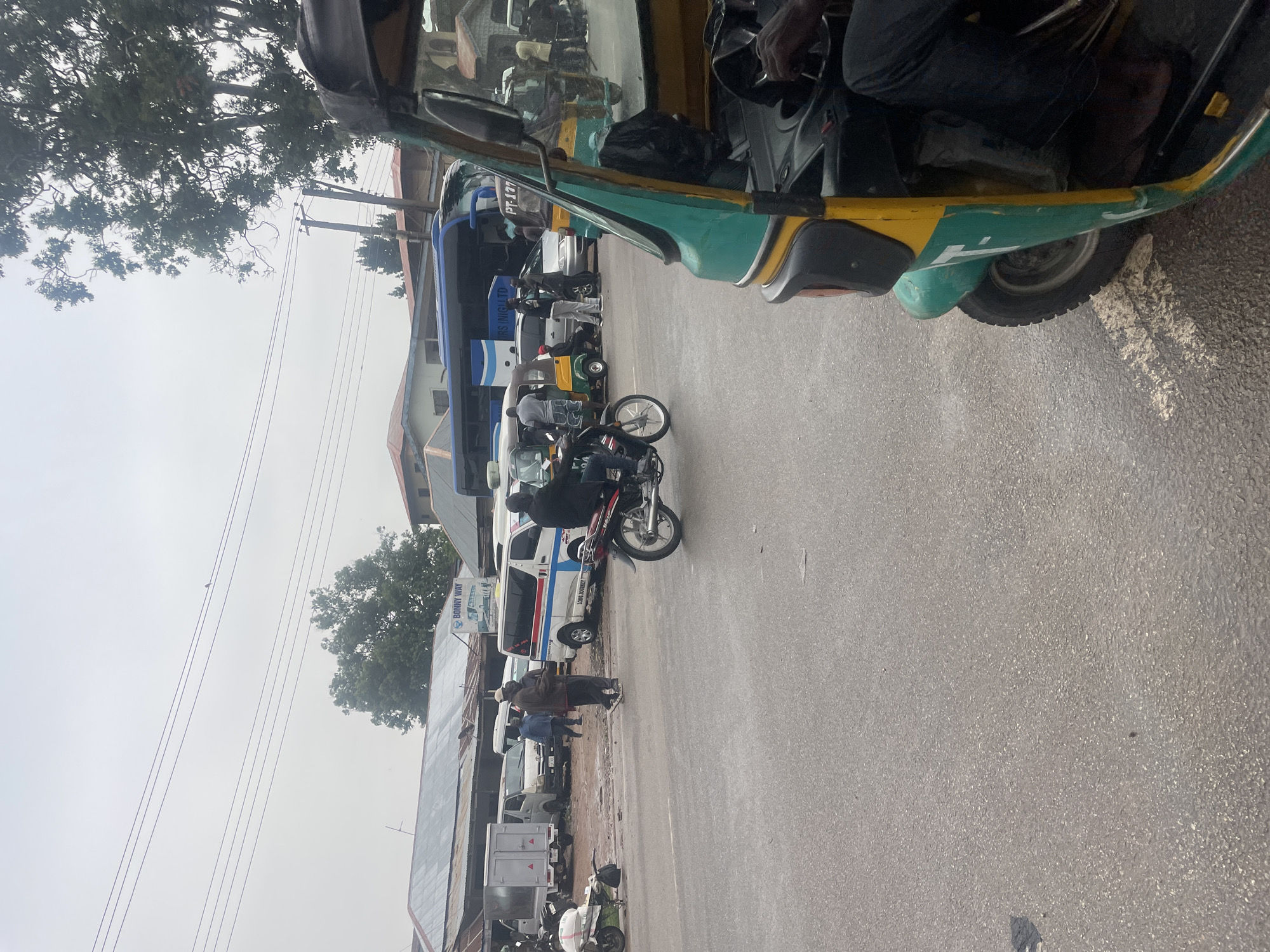 The image shows a street scene in Jos, Nigeria, with a busy transportation hub. In the foreground, a green and yellow auto-rickshaw is partially visible, with its driver's hand on the steering wheel. Behind it, a motorcycle with a rider is moving to the left. Further back, a variety of vehicles are parked and in motion, including vans, a large bus, and another auto-rickshaw. Several people are interacting with the vehicles, some appearing to be passengers or drivers. Buildings line the background, some with corrugated metal roofs, suggesting a mix of commercial and residential structures. The road surface appears wet, indicating recent rain. Overhead, power lines crisscross the sky, and trees with dense foliage are visible. The overall atmosphere is one of bustling urban activity.