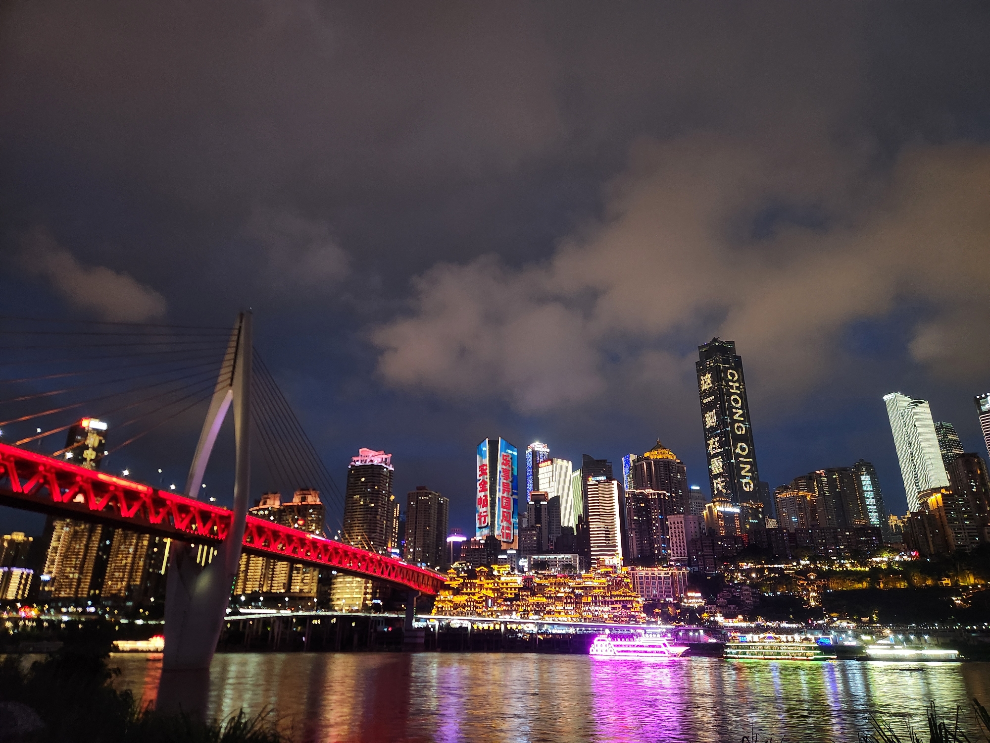Here's a description of the image:
The image depicts a nighttime cityscape dominated by illuminated skyscrapers and a cable-stayed bridge. The scene is located in 江北城街道, China. The sky is a dark gradient with patchy clouds.
A red bridge with white support cables stretches across the foreground, with reflections visible in the water. Numerous modern buildings, glowing with light, form the urban backdrop. Several buildings display vertical text or patterns of light. There are clusters of structures that resemble traditional Chinese architecture, also illuminated. Boats with bright lights are floating on the river. The water reflects the city lights in vibrant shades of pink, purple, and green. The bottom of the image shows some dark foliage.