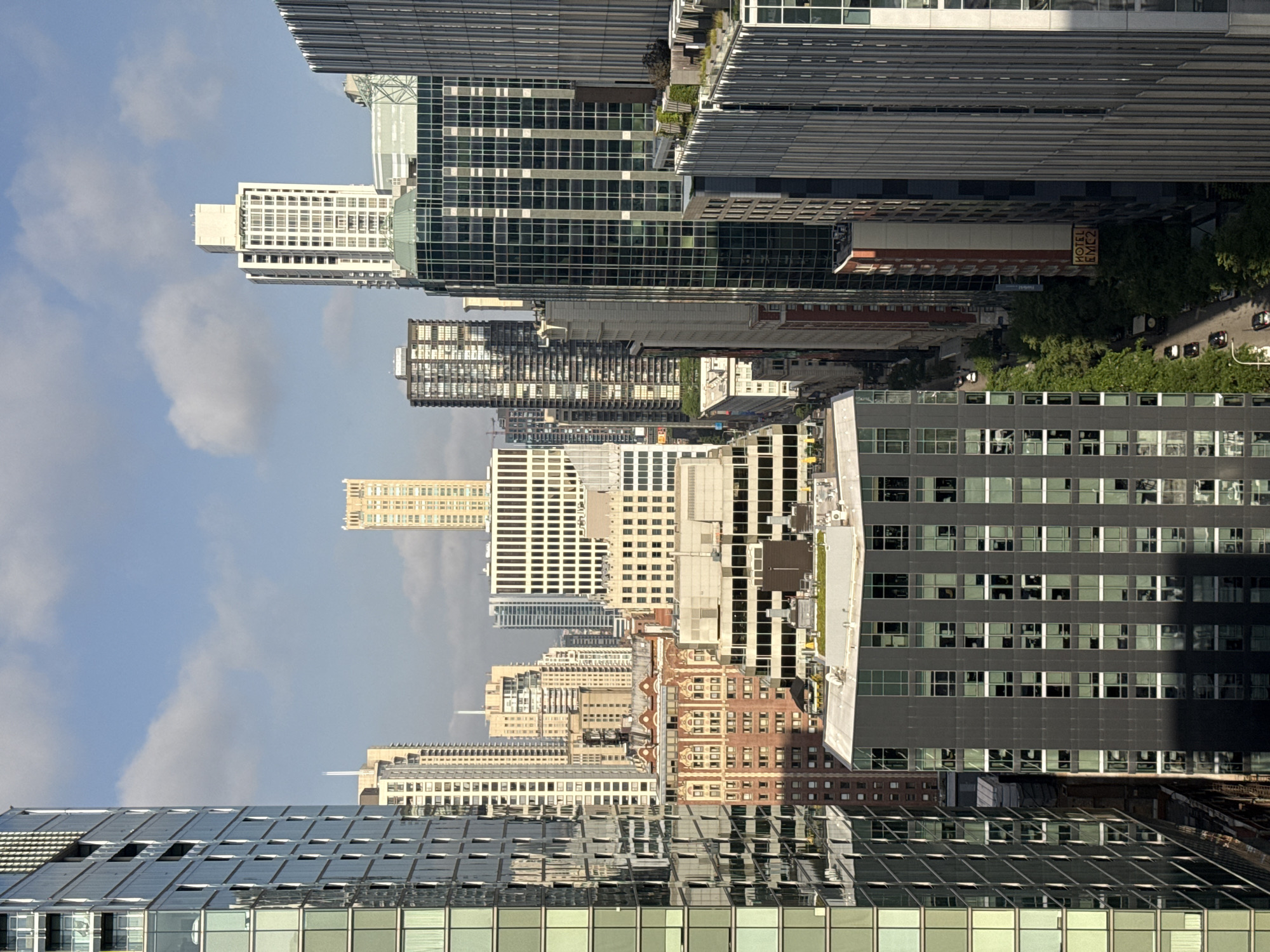 This media file depicts an urban cityscape with a focus on architecture. The scene is dominated by a dense collection of tall buildings, primarily skyscrapers, viewed from an elevated perspective. The buildings showcase a variety of architectural styles, from modern glass and steel structures to older brick buildings with more traditional window designs. Some buildings are clad in dark glass, reflecting the sky, while others are lighter in color with grid-like window patterns. Greenery is visible in pockets, suggesting parks or rooftop gardens, and trees line some of the streets.
The image appears to be taken during the daytime, likely late morning or afternoon, as indicated by the bright sky with scattered clouds and the shadows cast by the buildings. The lighting suggests fair weather. The orientation of the image is rotated 90 degrees clockwise.
Notable details include a sign on one of the buildings that reads "HOTEL EMC2". The sheer scale and density of the buildings imply an urban setting, and the reference to Chicago, United States of America, places this scene in a major metropolitan area known for its distinctive architecture. No people are clearly discernible in the image due to the distance and scale, and the focus is on the inanimate structures of the city. The overall mood is one of urban grandeur and the constant activity of a bustling city.