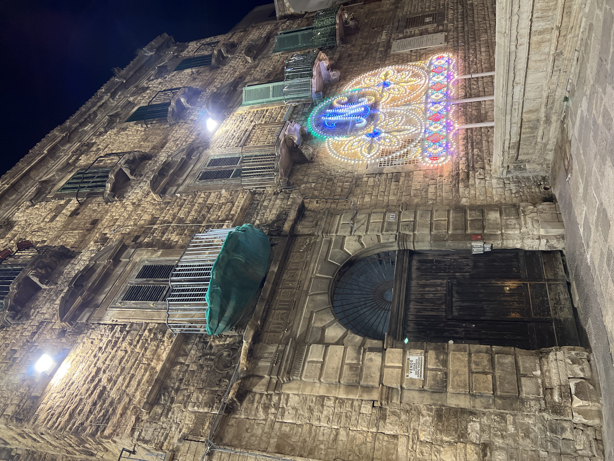 The image shows a nighttime street view of the exterior of a stone building in Bitonto, Italy. The building has multiple floors with balconies and shutters. There is a large arched wooden door at street level. A sign on the wall next to the door reads "VENDE LOCALE USO DEPOSITO DOPPIO INGRESSO". In front of the building, a large, illuminated decorative structure with intricate patterns and the letter "M" in blue is visible. The structure is made of white and colorful lights. The street in front of the building is paved with stone.