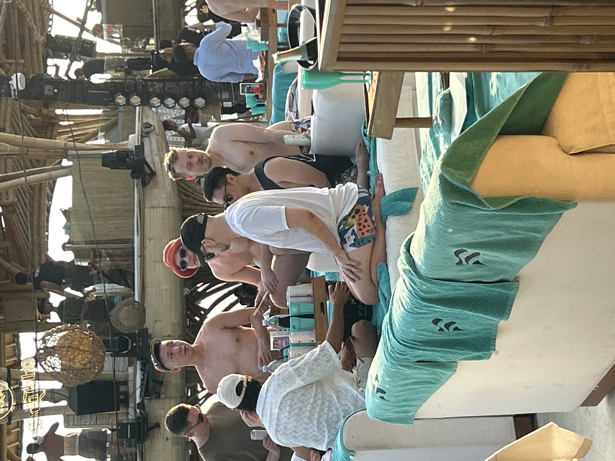 The image shows a group of men relaxing on loungers at what appears to be a beach club or resort in North Kuta, Indonesia. The setting features bamboo structures, open-air seating, and tropical foliage. In the foreground, several men are seated on a white sofa covered with teal towels. One man in a white patterned shirt and cap is facing away from the camera, engaged in conversation. Another man in colorful patterned shorts and a white t-shirt is also seated. Two men in the background, one wearing a red hat, are conversing. Beverages, including bottles and glasses, are visible on a table beside the loungers, suggesting a social gathering. The overall ambiance is relaxed and casual, typical of a vacation or leisure destination.