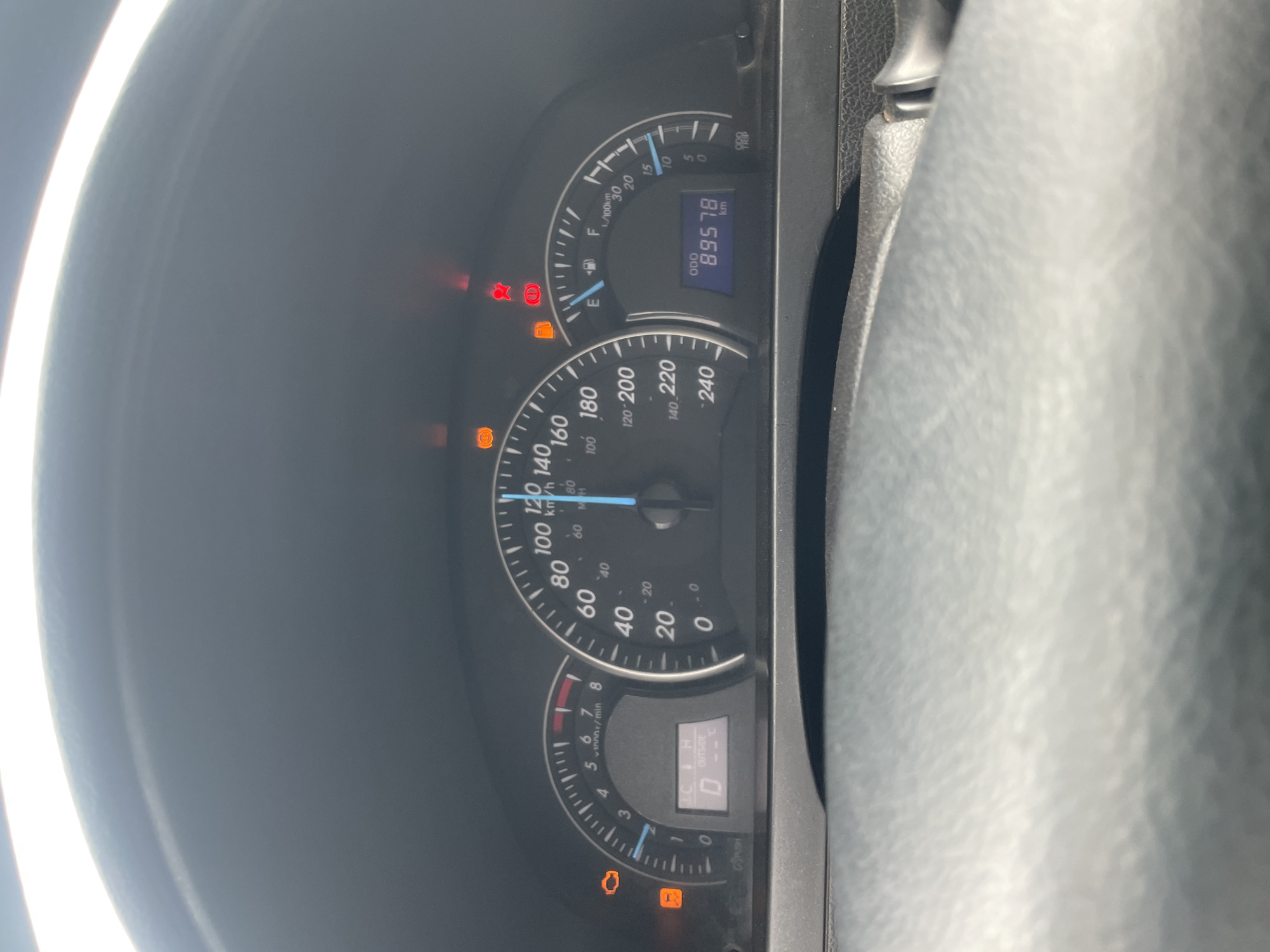 This is a close-up, low-angle, vertically oriented shot of a car's dashboard in Kwali, Nigeria. The main subjects are the car's instrument cluster, which includes a speedometer, a tachometer, a fuel gauge, and a digital display. The speedometer is marked in both km/h and mph, with the needle pointing to approximately 110 km/h. The tachometer is marked in rpm, with the needle near the 0 mark. The fuel gauge is showing nearly full. The digital display shows "ODO 89578 km," indicating the odometer reading. There are several warning lights illuminated on the dashboard, including what appears to be an engine light, a battery light, and a brake warning light. The ambient light suggests it is daytime. The overall scene is typical of a car's interior, with the focus on the functional elements of the dashboard.