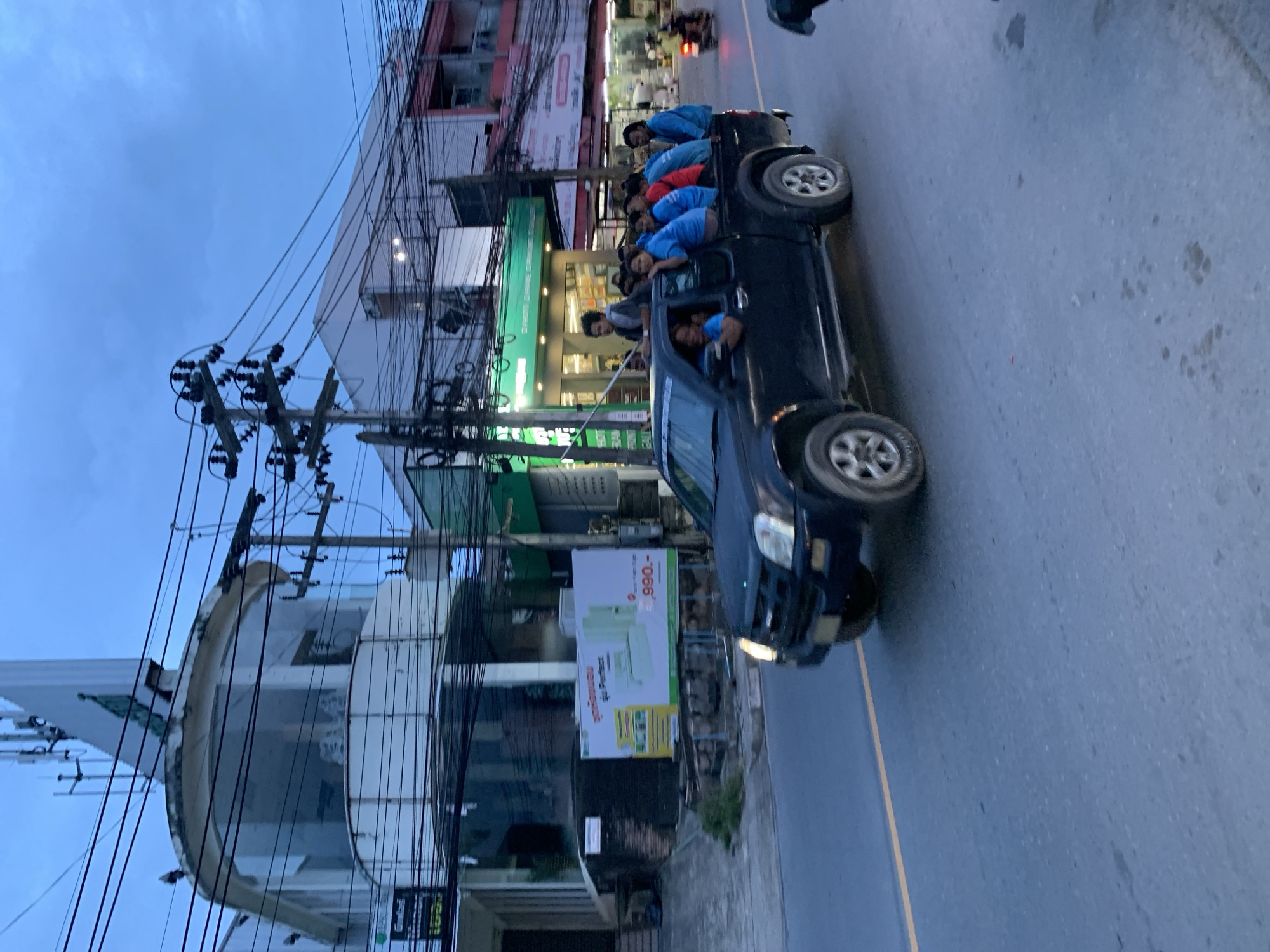 The image depicts a black pickup truck driving on a paved road in Baan Bo Phut, Thailand. Multiple people are riding in the back of the truck, some sitting and some leaning over the cab. Buildings line the street, and power lines are visible overhead. There's an advertising sign for a condominium with the price "990,-".