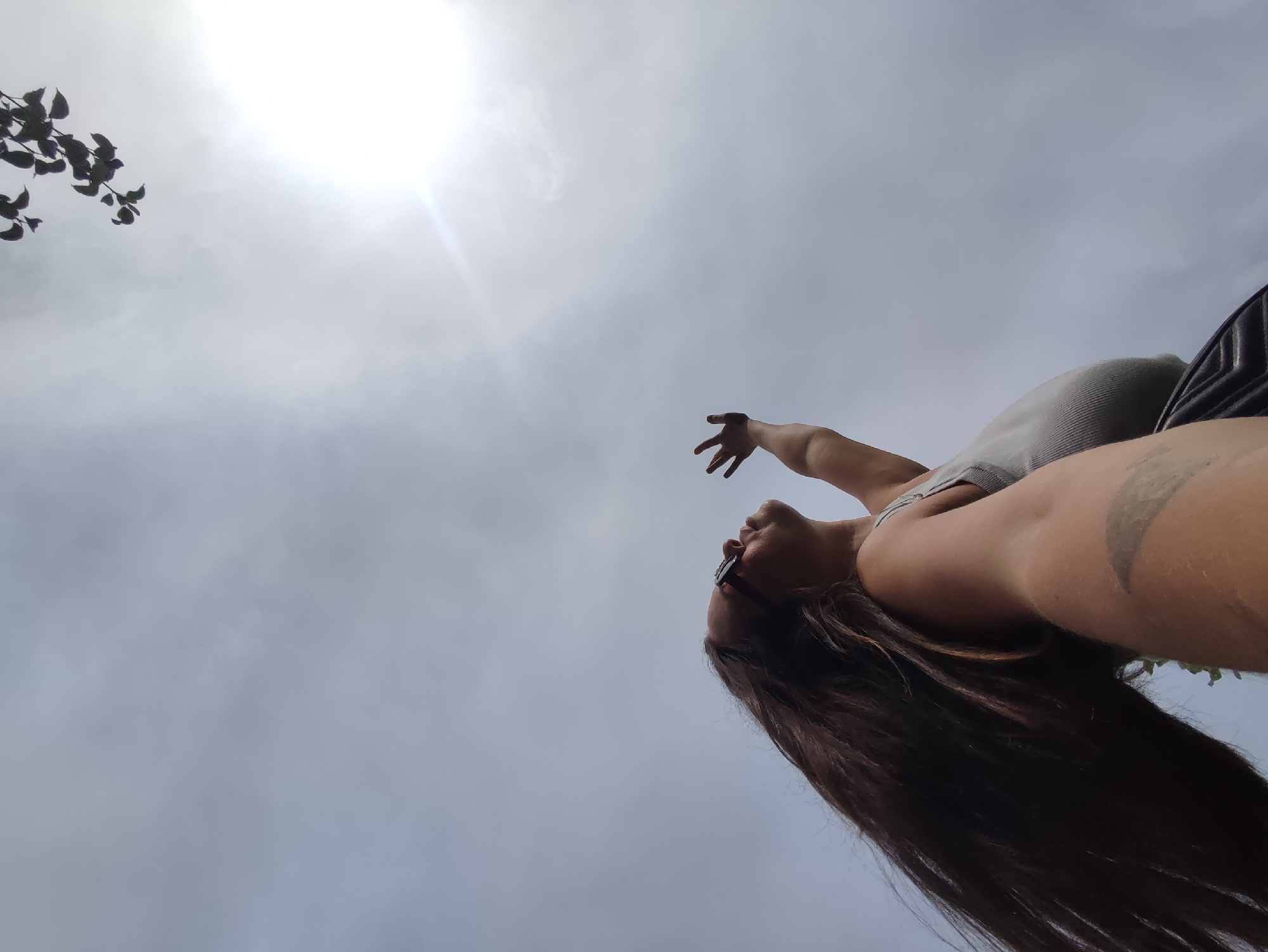 A low-angle shot captures a person viewed from below, with the sky filling most of the frame. The sun, a bright, diffused orb, is visible in the upper left quadrant, casting a gentle glow. To the left of the sun, a few dark leaves on a branch are silhouetted against the bright sky.
The individual, whose face is partially obscured and tilted upwards, appears to be wearing sunglasses and a grey tank top. Their long, dark hair cascades downwards, obscuring parts of their body. One arm is extended upwards and towards the right, with fingers splayed. Another arm is bent at the elbow, with a tattoo visible on the upper arm. The person seems to be in a relaxed or possibly celebratory pose.
The background is a hazy, overcast sky, with subtle variations in cloud cover. There is no indication of any specific urban environment. The scene does not provide any visual cues that would allow for the identification of its location within the city of Фоки, Russia.