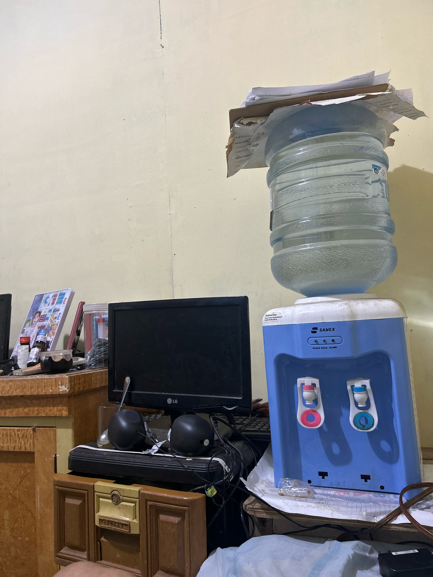 The image shows a cluttered desk area with a water dispenser in the foreground. The water dispenser is blue and white, with a large water bottle on top. There are two faucets on the front of the dispenser, one red and one blue. To the left of the water dispenser is a black computer monitor. On the desk are a pair of black speakers, a keyboard, and various papers and bottles. The wall behind the desk is a pale yellow color. The overall impression is one of a busy, functional workspace. The specific location in Sukanagara, Indonesia, is not discernible from the image alone.