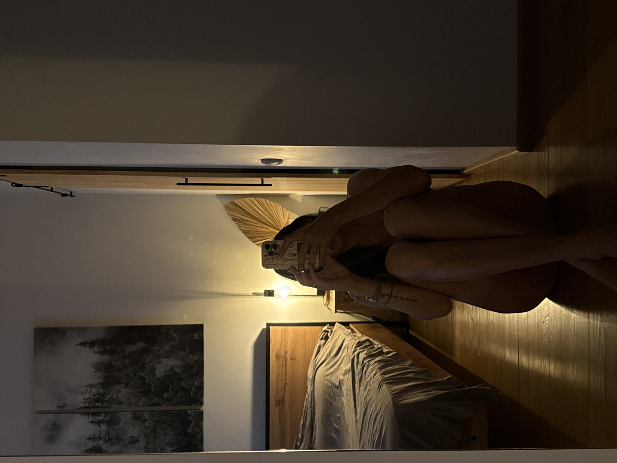 An adult, non-binary individual is sitting on a wooden floor in a dimly lit room, holding a phone to take a mirrored selfie. The individual is positioned in the foreground, facing a mirror that reflects their image. They are wearing minimal or no clothing, their body is obscured by their arms and the angle of the shot. Their legs are crossed and tucked towards their body. Tattoos are visible on their arms.
Behind the individual, a wooden bed with rumpled grey bedding is visible. To the left of the bed, two framed pieces of art depicting a misty forest scene hang on the wall. A bedside table with a lamp emitting a warm light stands near the bed. A decorative fan-shaped object is positioned on a wall behind the individual, partially visible in the reflection. A wooden door with a sliding track is to the right of the frame. The floor is polished wood, reflecting the light from the lamp. The environment suggests a bedroom or a similarly intimate personal space. There is no indication of the location being in the city of Chaikovsky, Russia.