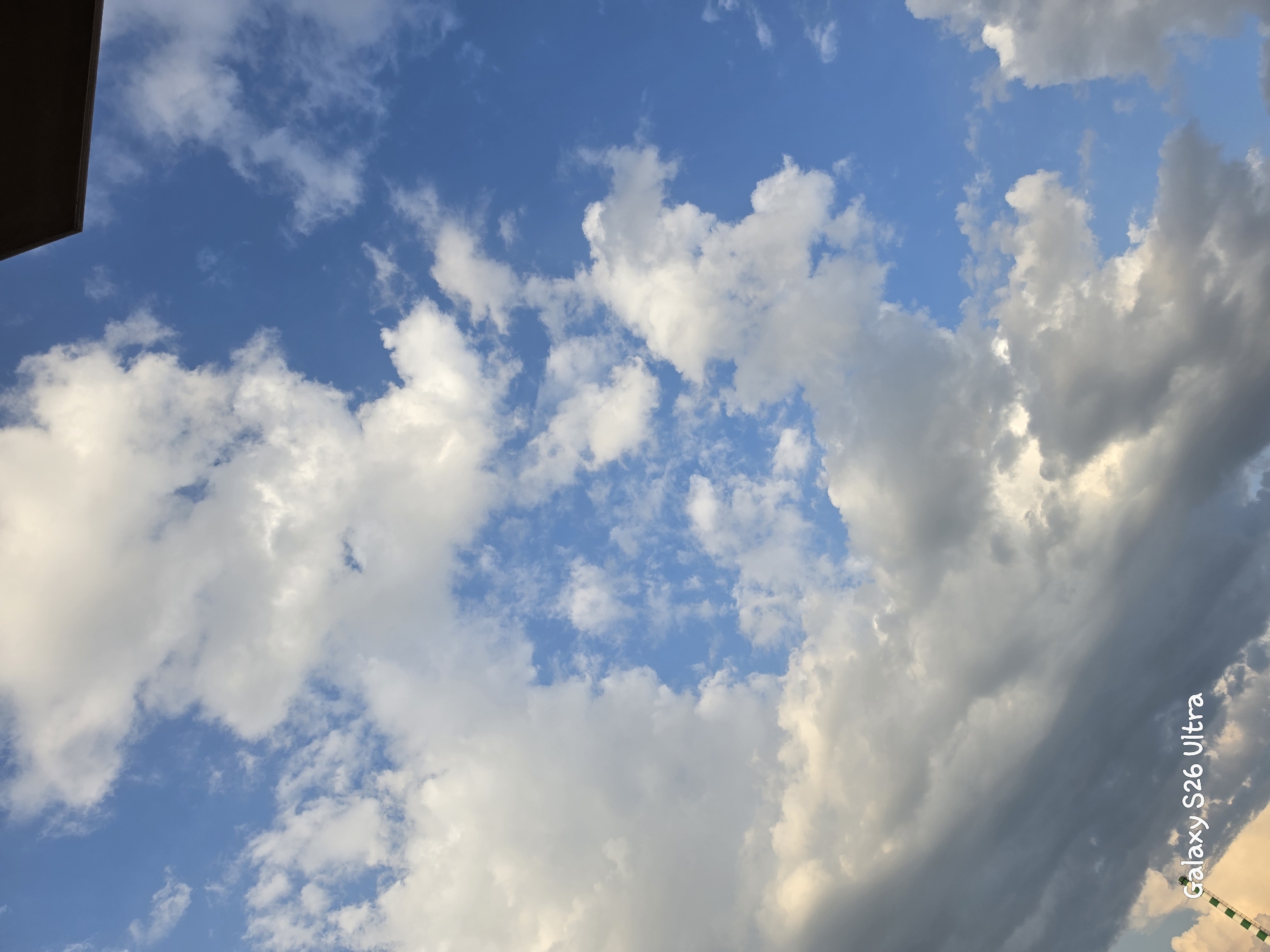 The image captures a dynamic sky filled with a mix of fluffy white cumulus clouds and darker, more ominous storm clouds. The bright blue sky peeks through in patches, suggesting a transition in weather. Sunlight illuminates some of the clouds from the side, creating highlights and shadows, hinting at a late afternoon or early evening.
In the upper left corner, a dark, angular object partially enters the frame, possibly the edge of a building or roof, providing a subtle hint of an urban or constructed environment. The overall scene is dominated by the sky, conveying a sense of vastness and the power of nature. There are no people or explicit human activity depicted.
The visible text "Galaxy S26 Ultra" is watermarked diagonally on the right side of the image, indicating it is a photograph taken with a Samsung Galaxy S26 Ultra phone.
The location context of Plovdiv, Bulgaria, suggests this sky is viewed from that region. The weather appears to be unsettled, with a storm potentially approaching or passing, characterized by the dramatic contrast between light and dark clouds.
