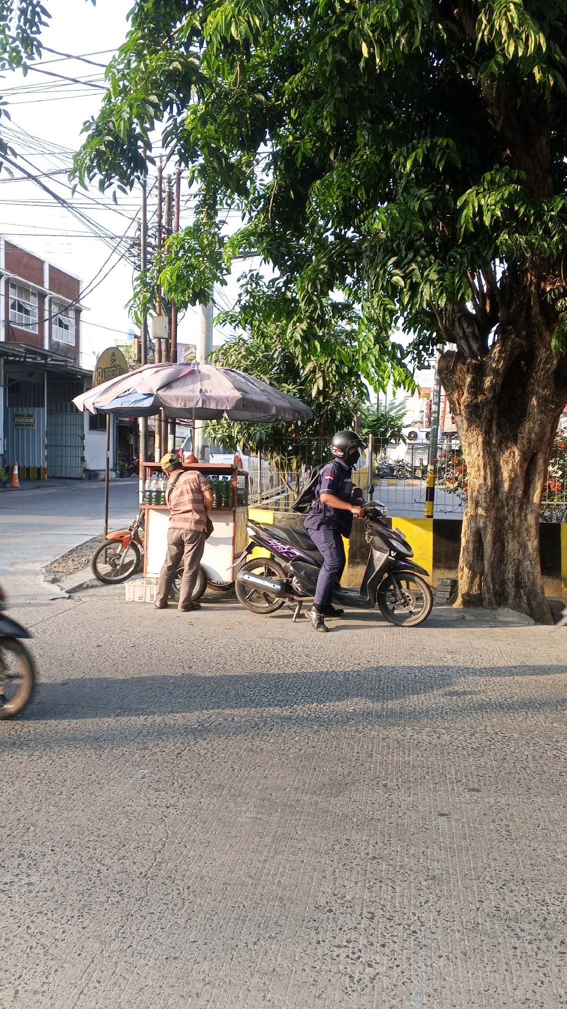 The image depicts a street scene in Semarang, Indonesia. In the foreground, a person wearing a dark blue uniform and a black helmet is seated on a dark-colored scooter. The scooter is parked on the asphalt road, which exhibits a coarse texture and parallel grooving. To the left of the scooter, a vendor is standing behind a small mobile cart that is partially covered by a faded umbrella. The cart displays several bottles of liquid, likely beverages, arranged on shelves. Another scooter, orange in color, is parked to the left of the vendor's cart. In the background, a large tree with dense green foliage dominates the right side of the frame. Behind the vendor's cart, a building with a brown facade and white window frames is visible. The scene is illuminated by natural light, casting shadows on the road. Overhead, electrical wires crisscross the sky.
