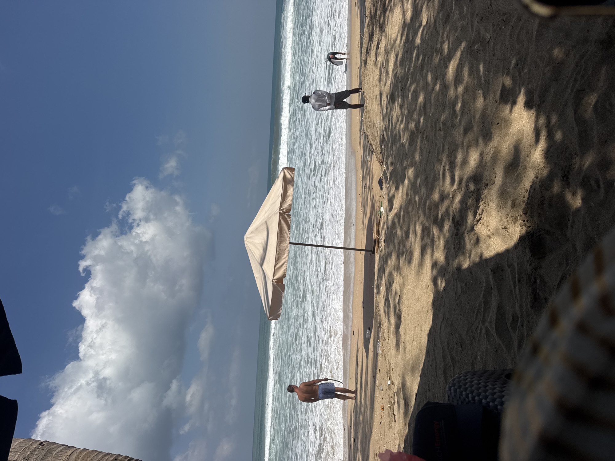 This image depicts a sunny day at Kuta Beach, Bali, Indonesia. The foreground is occupied by the sandy beach with scattered debris and the shadows of palm trees. A black baseball cap rests on a woven chair in the lower left corner. Further back, an open umbrella shades a patch of sand. Two men are visible on the shoreline. One man, wearing blue shorts, stands facing the ocean, holding a dark object. Another man, dressed in a white shirt and grey shorts, faces away from the camera, looking towards the water where a third person, partially obscured, appears to be interacting with the waves or sand. The ocean is active with moderate waves breaking near the shore. The sky is blue with scattered white clouds.