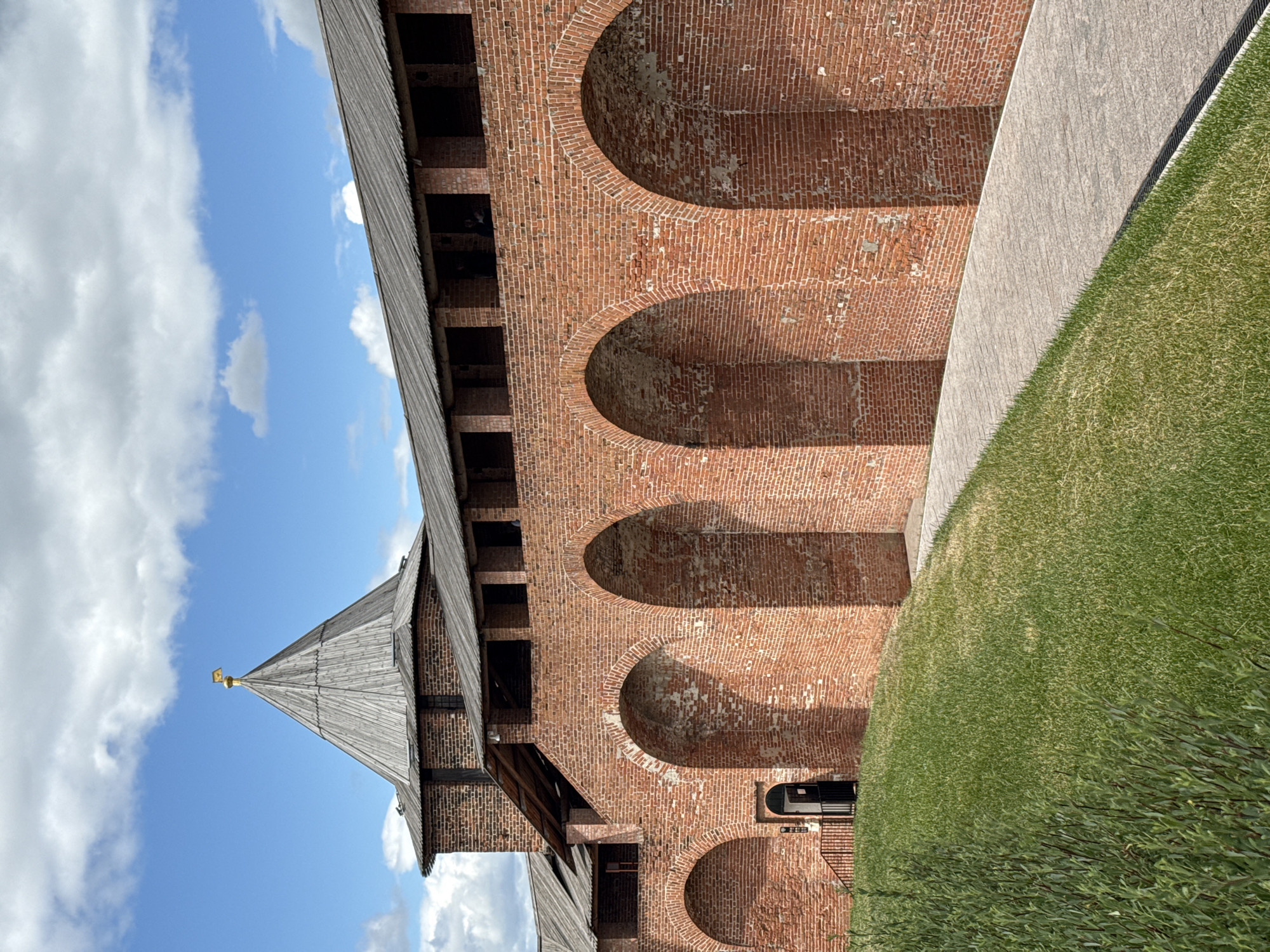 The image shows a view of the Nizhny Novgorod Kremlin, specifically the Wall and the Taynitskaya Tower. The wall, constructed of red brick, features a series of arched openings and is topped with a wooden roof. The Taynitskaya Tower, also made of brick, has a conical wooden roof with a golden finial. A paved pathway runs alongside the wall, bordered by a grassy slope and some low-lying vegetation. The sky is blue with scattered white clouds. No people are clearly visible in the scene. The lighting suggests it is daytime, likely with the sun out.