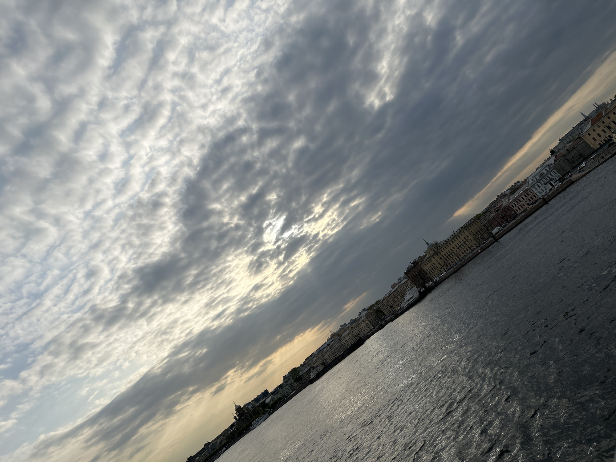 The image is a tilted, eye-level view of the Neva River in Saint Petersburg, Russia. A body of water, the Neva, covers approximately half the bottom portion of the image, the water's surface reflecting the light of the sky. Parallel to the river lies a cityscape of classical architecture, featuring buildings with ornate facades in varying shades of beige, cream, and light brown. Some of the buildings are multi-storied. The upper half of the image displays a cloudy sky, with a mix of gray, white, and dark gray clouds. The lighting is diffused, with the sun obscured by the cloud cover, resulting in a soft and somewhat muted atmosphere. The image is tilted to the right.
