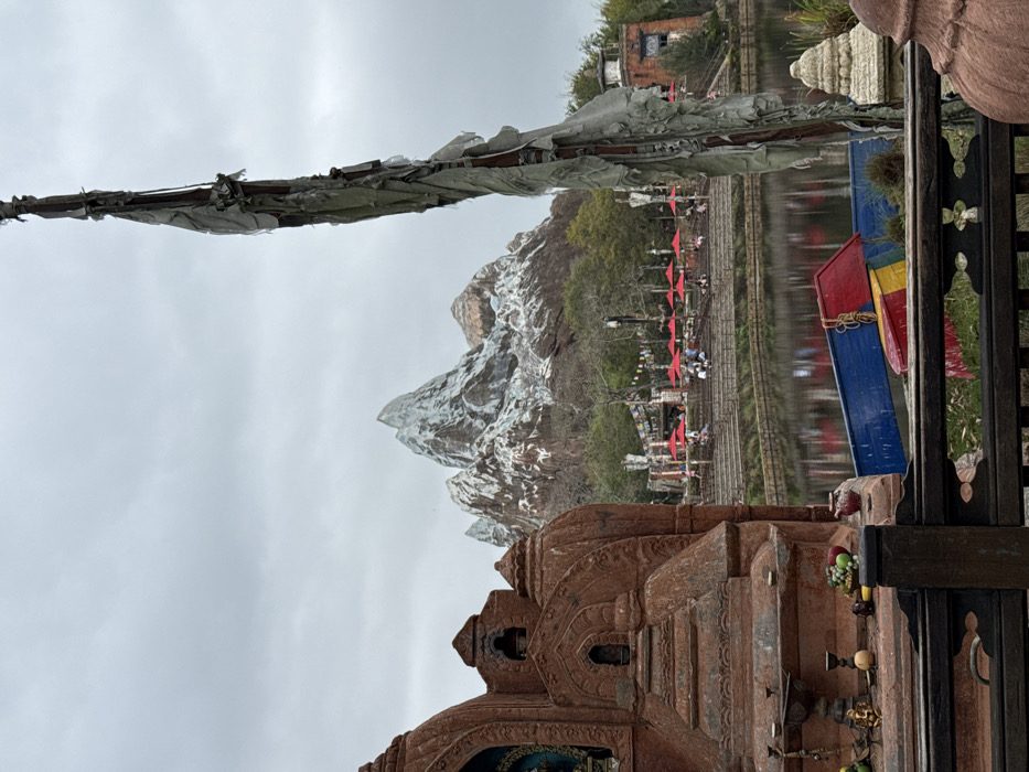 The image depicts an outdoor scene in the city of Bay Lake, Florida, specifically within Disney's Animal Kingdom theme park, focused on the Asia section featuring Expedition Everest. An overcast sky dominates the upper portion of the frame. In the left foreground stands a large, multi-tiered reddish-brown stone structure resembling a shrine, featuring intricate carvings, arched niches (one containing a blue and gold figure), and a flat surface displaying offerings including small fruits, a brass vessel, and a red pot. A dark wooden railing with decorative cutouts crosses the bottom foreground. To the right, a tall, weathered wooden pole is draped with numerous tattered, faded gray fabric strips.
A calm body of water spans the midground, reflecting the sky and distant elements. Three small, brightly colored boats (blue, red, yellow/green) are partially visible at the water's edge, seemingly overturned or resting. Beyond the water, a terraced area with stone steps leads to an outdoor dining or rest area. Multiple individuals are present, seated at tables under numerous red patio umbrellas. Colorful prayer flags are strung between posts in this area. In the far background, a large, artificial snow-capped mountain range, identifiable as Expedition Everest, rises prominently against the gray sky, flanked by trees at its base.