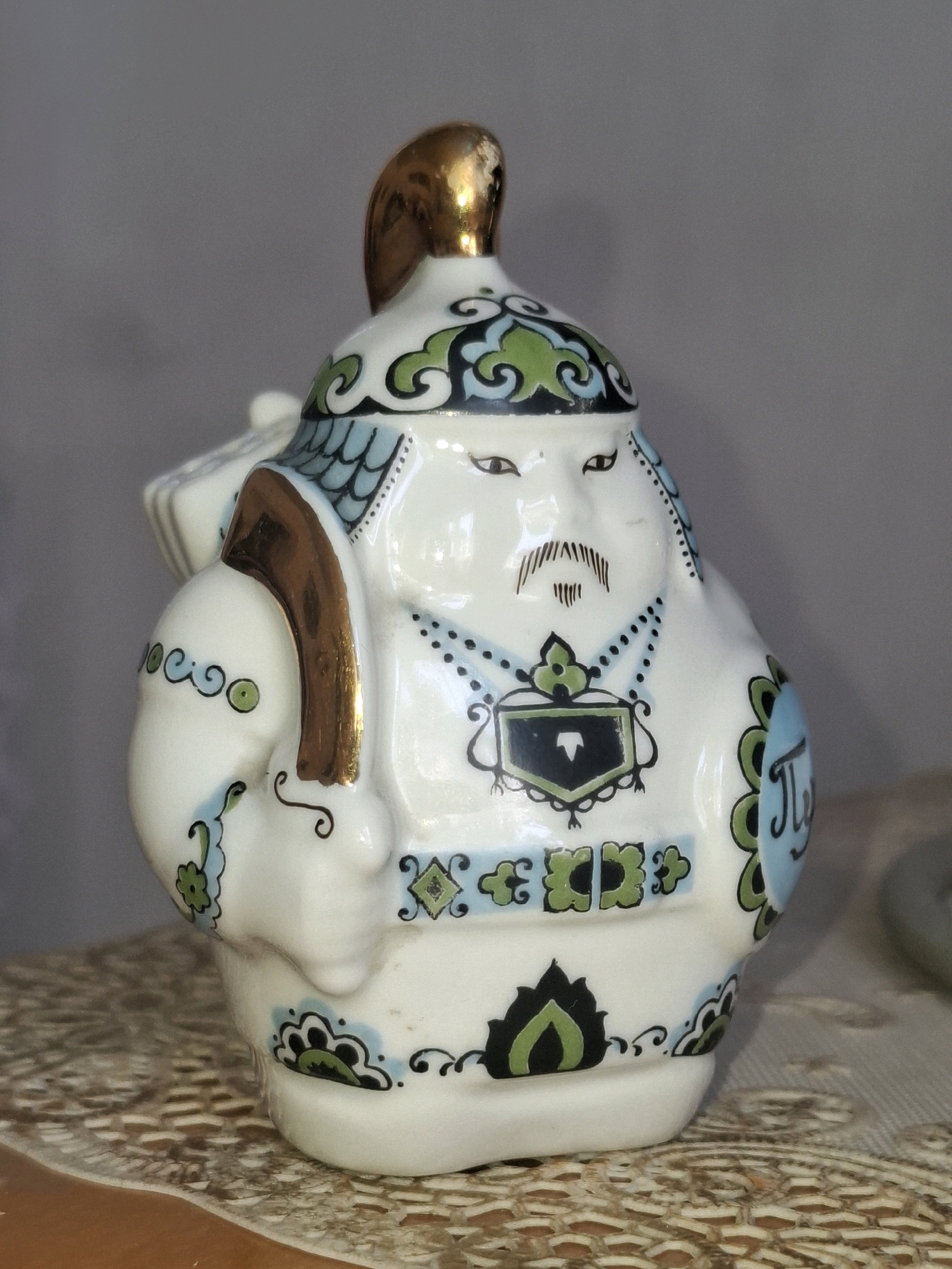 A close-up, eye-level studio shot shows a small, white, ceramic figurine of a stout, stylized man. The figurine is decorated with intricate patterns in blue, green, and black, with gold accents on the helmet crest and a shoulder strap. The man has a painted face with wide eyes, a mustache, and a goatee. He appears to be wearing a helmet and possibly carrying something on his back. The figurine is placed on a textured, light-colored surface, likely a doily or placemat, which rests on a wooden surface. The background is a plain, light gray wall. There is no indication of the city of Almaty, Kazakhstan, or any specific urban environment.