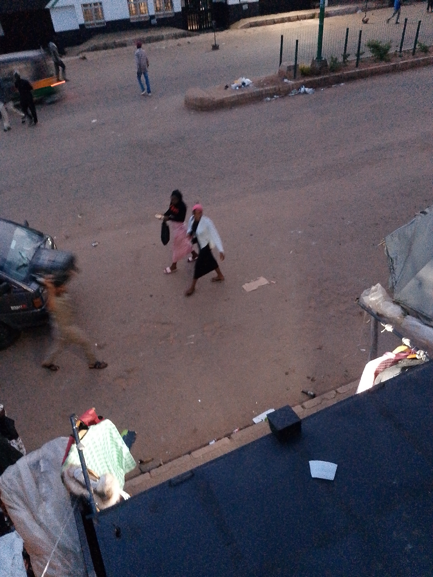 The image depicts an outdoor scene, likely a street or sidewalk in Bukuru, Nigeria, during the evening or dusk. The lighting is dim, contributing to the grainy quality of the photograph.
In the foreground, a dark, flat surface, possibly the roof of a building or a vehicle, occupies the bottom right portion of the frame. A small, light-colored rectangular object, possibly a piece of paper or debris, is visible on this surface. To the left of this dark surface, a cluttered area shows various items, including fabric, possibly clothing or bedding, and a light-colored tent-like structure. A black cube-shaped object sits on the edge of the paved area.
The middle ground features a paved surface, likely a road or pathway. Several people are present. To the left, a dark-colored car is visible, with what appears to be a person, blurred by motion, carrying a large object over their shoulder. Further to the right, two women are walking. One, wearing a pink skirt and black top, carries a dark bag. The other wears a white jacket and a pink headscarf.
In the background, more people are seen walking. One individual in a blue top and jeans stands further down the paved area. The backdrop includes a building with multiple windows, suggesting it is a commercial or residential structure. A green vertical pole is also visible. The ground surface appears to be a mix of asphalt and dirt. Debris is scattered along a low wall or curb. The overall impression is of a busy, everyday scene in a Nigerian city at the end of the day.
