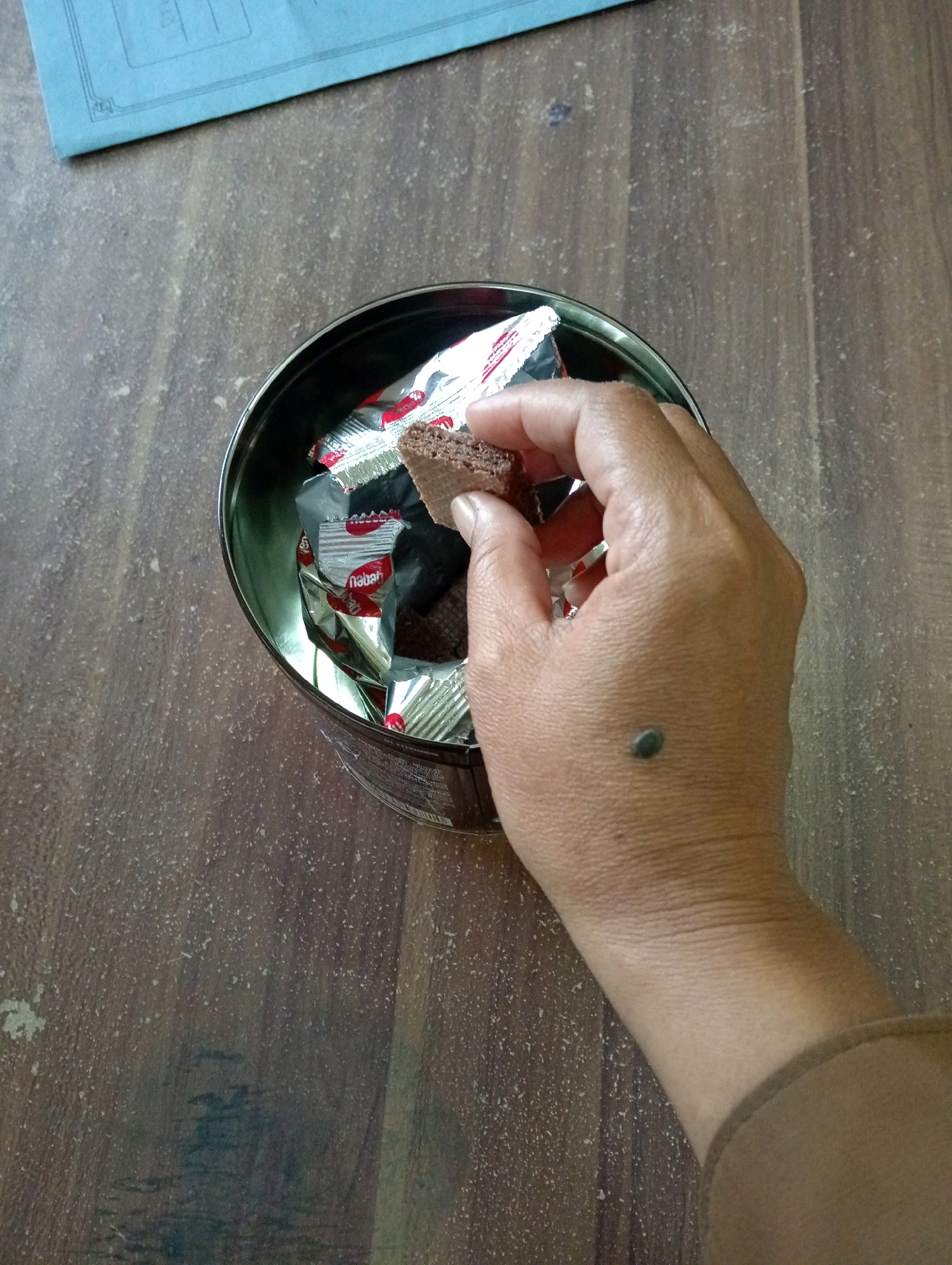 An overhead shot shows a hand holding a chocolate wafer cookie over a tin filled with individually wrapped chocolate wafer cookies. The hand has a dark mole on the back. The tin is sitting on a wooden surface, and a blue folder is partially visible in the top left corner. The overall scene suggests a casual, indoor setting, possibly a home or office break room. There is no indication of a specific location in Sidoharjo, Indonesia.