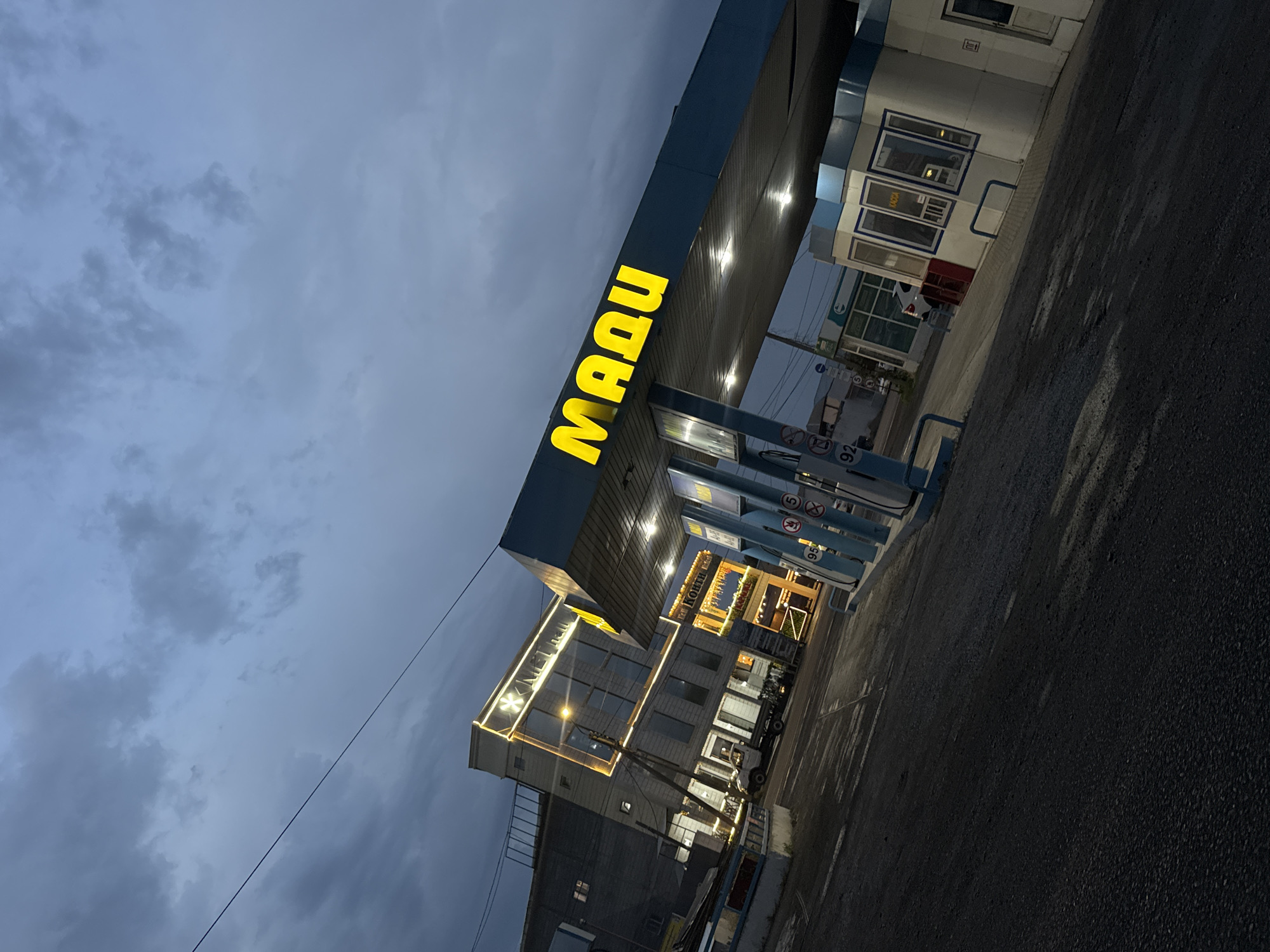 The image depicts a gas station in Taraz city administration, Kazakhstan, at dusk. The sky is cloudy and dark blue. A blue awning with yellow illuminated letters spelling "MAQV" is prominent. Beneath the awning are gas pumps, one of which has a "92" indicator. To the left, a multi-story building is illuminated with yellow lights outlining its windows and a sign that appears to read "METAN". Various vehicles, including a white truck and a white car, are visible. The ground is asphalt, appearing wet and reflecting the lights. Other structures, possibly shops or offices, are in the background. The overall scene suggests a commercial area at twilight.