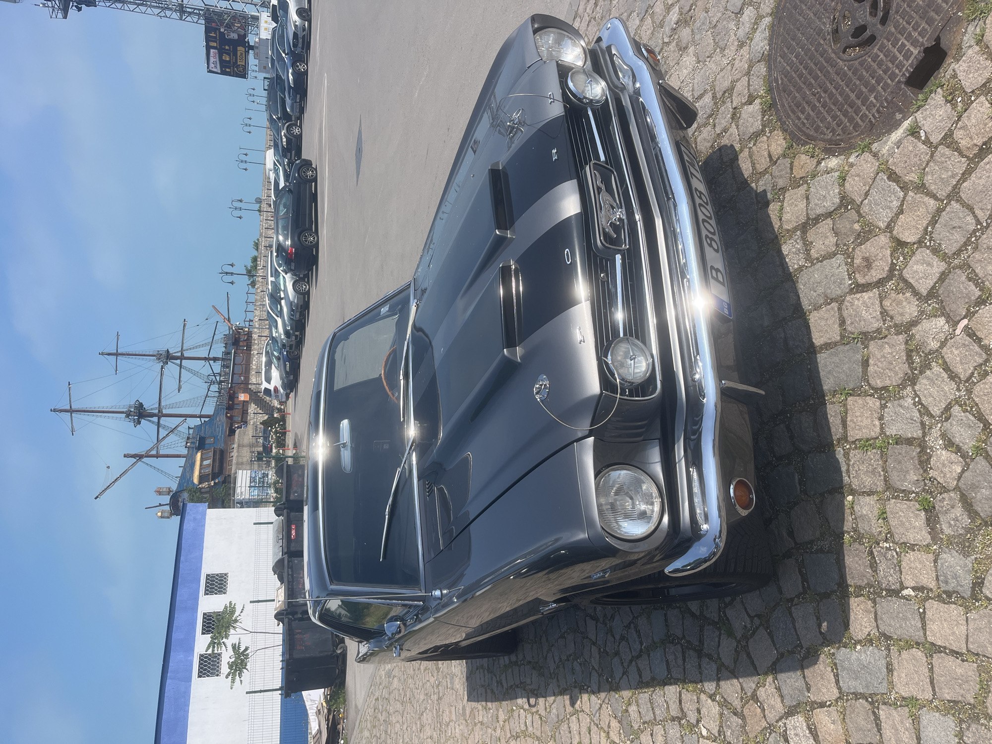 This media file captures a classic dark grey Ford Mustang convertible, likely a first-generation model from the mid-1960s, parked on a cobblestone surface in Varna, Bulgaria. The car, featuring two prominent lighter grey racing stripes along its hood, showcases chrome detailing on its grille, bumpers, and headlight bezels. A glimpse of the interior reveals a wooden steering wheel. The front license plate, partially obscured by sun glare, displays "B 8008 TT" and the "BG" country code, confirming its Bulgarian registration.
The car is situated outdoors on a bright, sunny day, evidenced by the clear blue sky and strong shadows cast on the ground, suggesting midday or early afternoon. The ground transitions from irregular cobblestones near the Mustang to an asphalt parking area for a row of modern, dark-colored cars in the background. To the left, a large, multi-masted wooden sailing ship is docked, indicating a harbor or port environment. Behind the ship, a white building with blue trim and grilled windows is visible.
In the upper background, a sign written in Bulgarian can be partially seen, displaying "бърза храна" (fast food) and "150м," suggesting a nearby eatery. A metal manhole cover is visible on the cobblestones beside the car. The scene is devoid of people, presenting a calm, static snapshot of the classic vehicle against the backdrop of a historic port city.