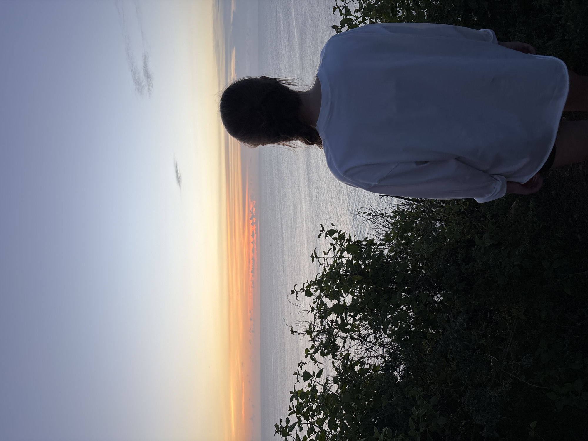 A woman with dark hair is standing on a cliff in Pecatu, Indonesia. She is facing away from the camera, overlooking the ocean. She is wearing a white long-sleeved shirt. The sky is a gradient of colors from orange to blue, suggesting a sunrise or sunset. The ocean is calm and reflective. The cliff is covered in green vegetation.