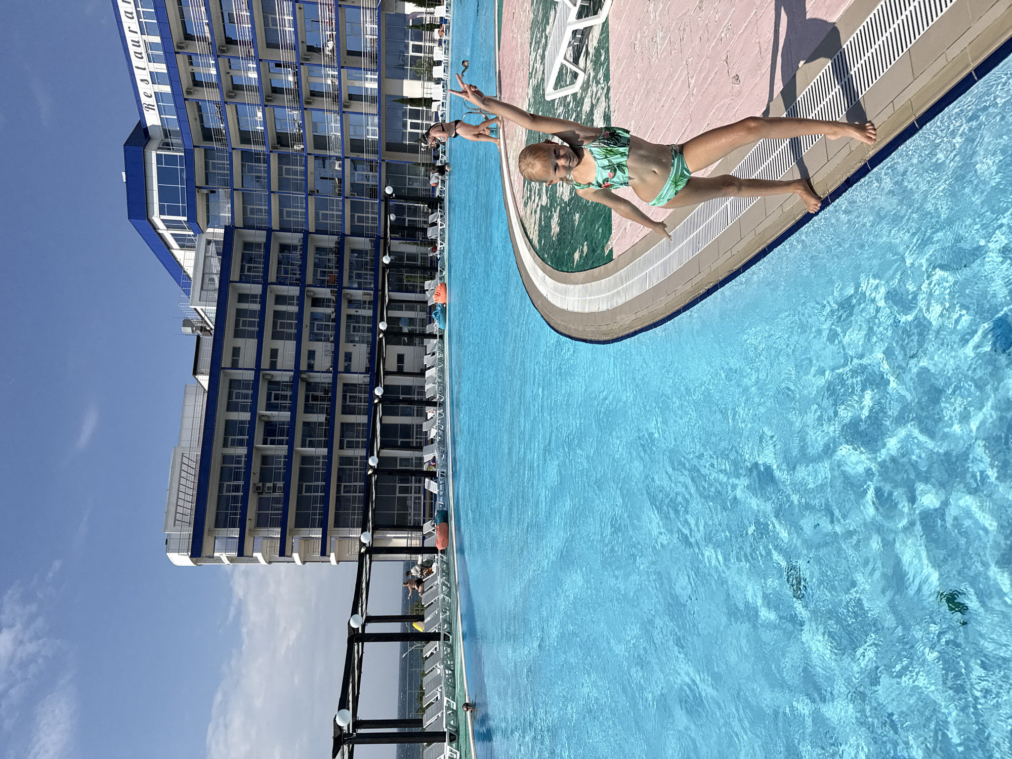 A young girl in a green swimsuit with a tropical pattern is posed playfully at the edge of a bright blue swimming pool. She has her arms outstretched, as if she is about to jump or has just emerged from the water. Her expression is joyful. Behind her, another person, possibly a child, is also in the pool, wearing goggles and a swimming cap. The pool area is bordered by a pink and green tiled edge, with white lounge chairs visible in the background. To the left of the pool, a tall, modern blue and white building stands under a clear blue sky with scattered clouds. The building has many windows and balconies, and text reading "Restauran" is visible on its side. The scene appears to be set on a sunny day, likely during daylight hours, suggesting a vacation or leisure setting. The orientation of the image is landscape, with the top of the building on the left and the pool occupying the right side.