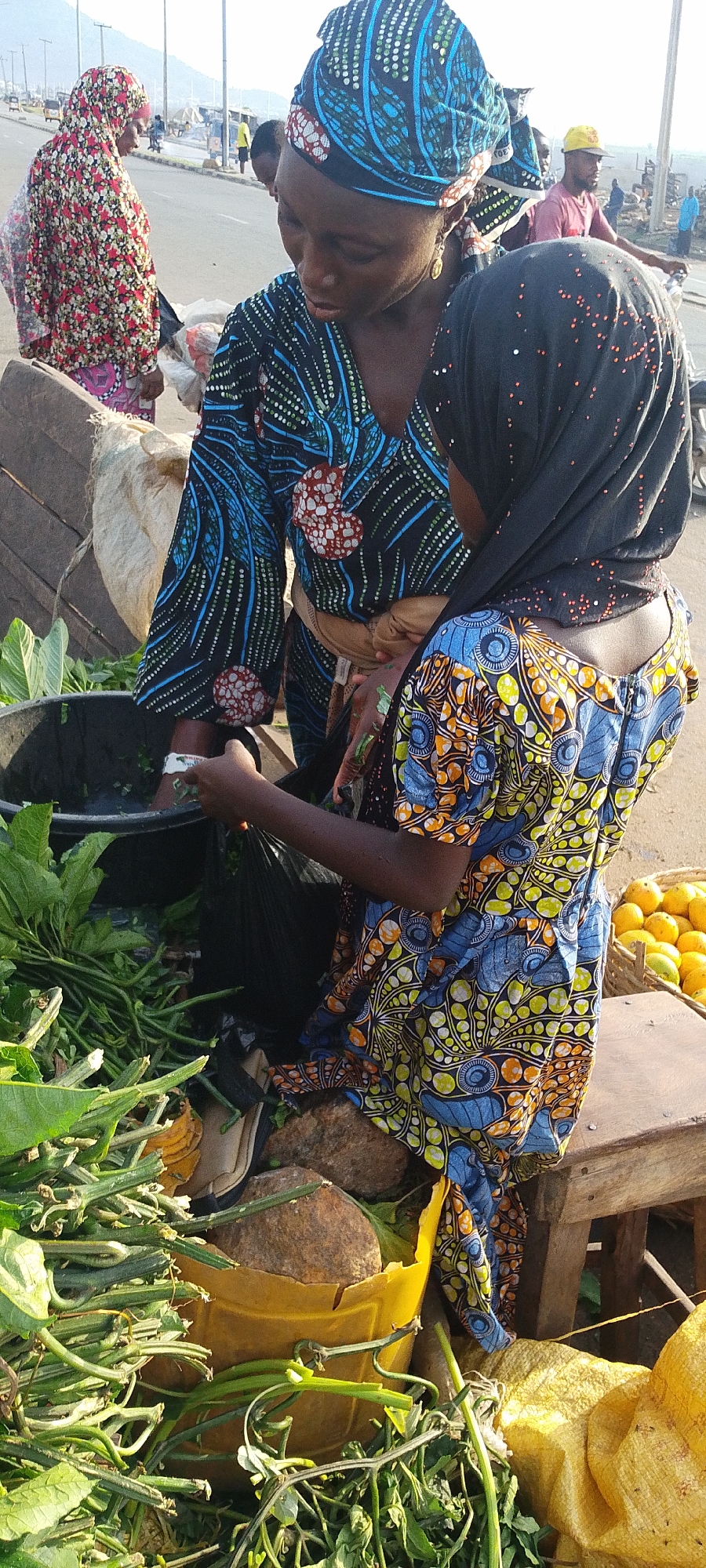 In Lokoja, Nigeria, two women are engaged in a transaction at an outdoor market stall. The woman on the left, wearing a blue and black patterned headwrap and a similarly patterned dress, appears to be selling leafy green vegetables. She is holding a handful of the vegetables. The other woman, standing to the right, is wearing a black headscarf with small red dots and a blue and yellow patterned dress. She is holding a black plastic bag. Between them is a black plastic container filled with water and leafy greens, along with a pile of the same vegetables extending onto the ground. A yellow bucket and various other items are nearby, and a basket of oranges can be seen to the right. In the background, there are people and vehicular traffic on a road, with a mountain visible in the distance.