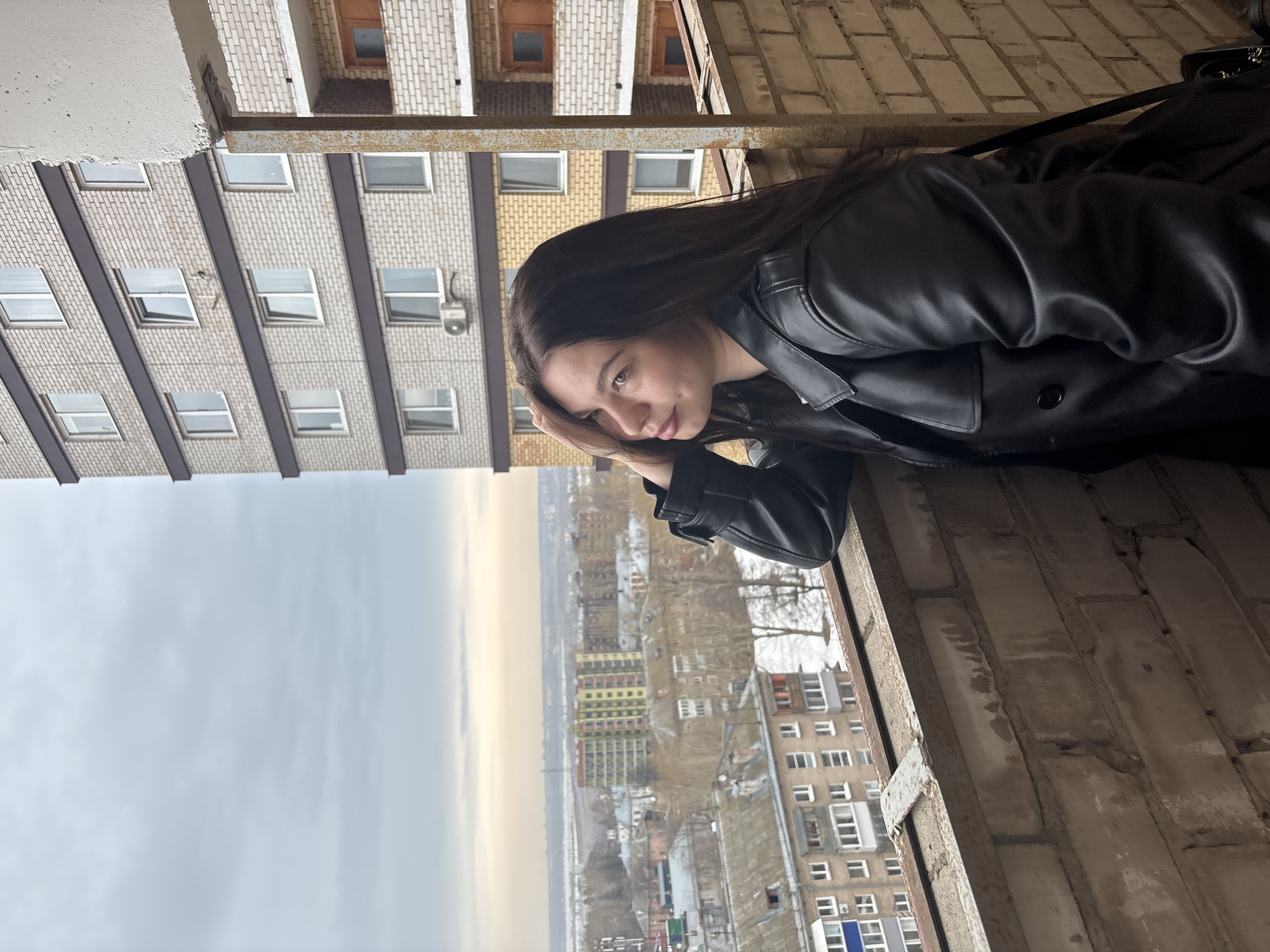 A young woman with long, dark hair, wearing a black leather jacket, leans against a brick windowsill. Her left hand is supporting her head, and she gazes out over a city landscape. The photograph appears to have been taken from a high vantage point, possibly a balcony or high window, overlooking a densely populated urban area. Buildings of various heights and architectural styles fill the scene. The sky is overcast and gray, suggesting a cloudy day.
Based on the architectural style of the buildings and the urban layout, the location can be identified as Perm, Russia.