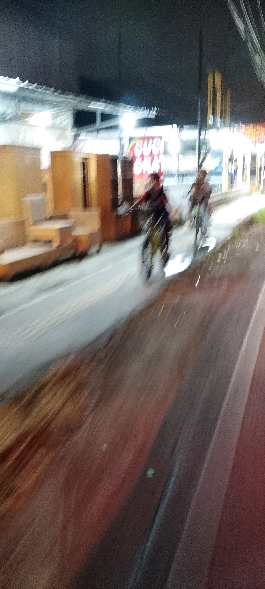 The image captures a blurry night scene on a street in Karangsatria, Indonesia. Two individuals are riding motorcycles, one in the foreground and another slightly behind and to the right. The person in the foreground is wearing dark clothing and appears to be turning their head to the left. The motorcycle in the foreground has visible spokes on the wheels. Behind the motorcycles, there are structures that resemble stalls or shops, with some bearing text that is difficult to decipher due to the blurriness, but parts of "SUC" and "GRA" are discernible on a red sign. The street appears wet, reflecting the lights from the surroundings, and the motion blur suggests movement. The overall environment is dark with artificial lighting, typical of a commercial or residential area at night.