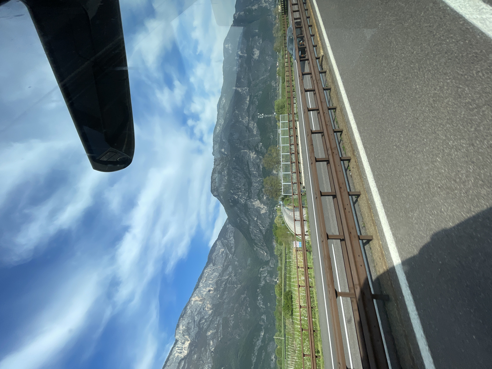The image, taken from inside a vehicle, shows a view of Zambana, Italy, through the windshield. A section of the vehicle's rearview mirror is visible on the left. Outside, a mountain range dominates the landscape. To the right, a multi-lane highway runs alongside a green area, possibly vineyards, separated by fencing and barriers. The sky is blue with scattered clouds.