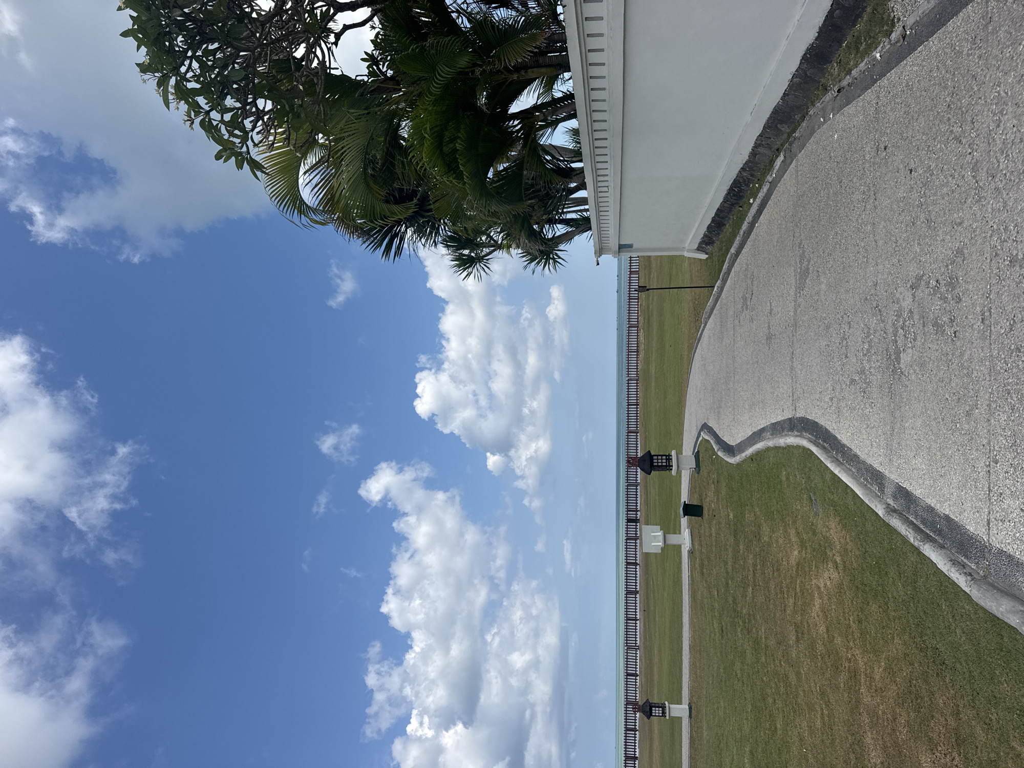 The image depicts a pathway leading towards the sea in Kuta Selatan, Indonesia. The pathway is paved with a speckled gray material and is bordered by a strip of grass on the left and a white wall on the right. The white wall is topped with a decorative railing and is partially obscured by lush green palm trees and other tropical foliage. Beyond the grass, a dark wooden fence runs along the coastline, separating the manicured lawn from the turquoise sea. The sea is calm, with a hazy horizon. The sky above is a vibrant blue, dotted with fluffy white clouds. Two ornate, dark-colored lanterns on white pedestals are visible on the lawn, along with a green trash bin and a white structure that might be a signpost or a small monument. The overall scene suggests a resort or a well-maintained public space near the beach, bathed in bright daylight.