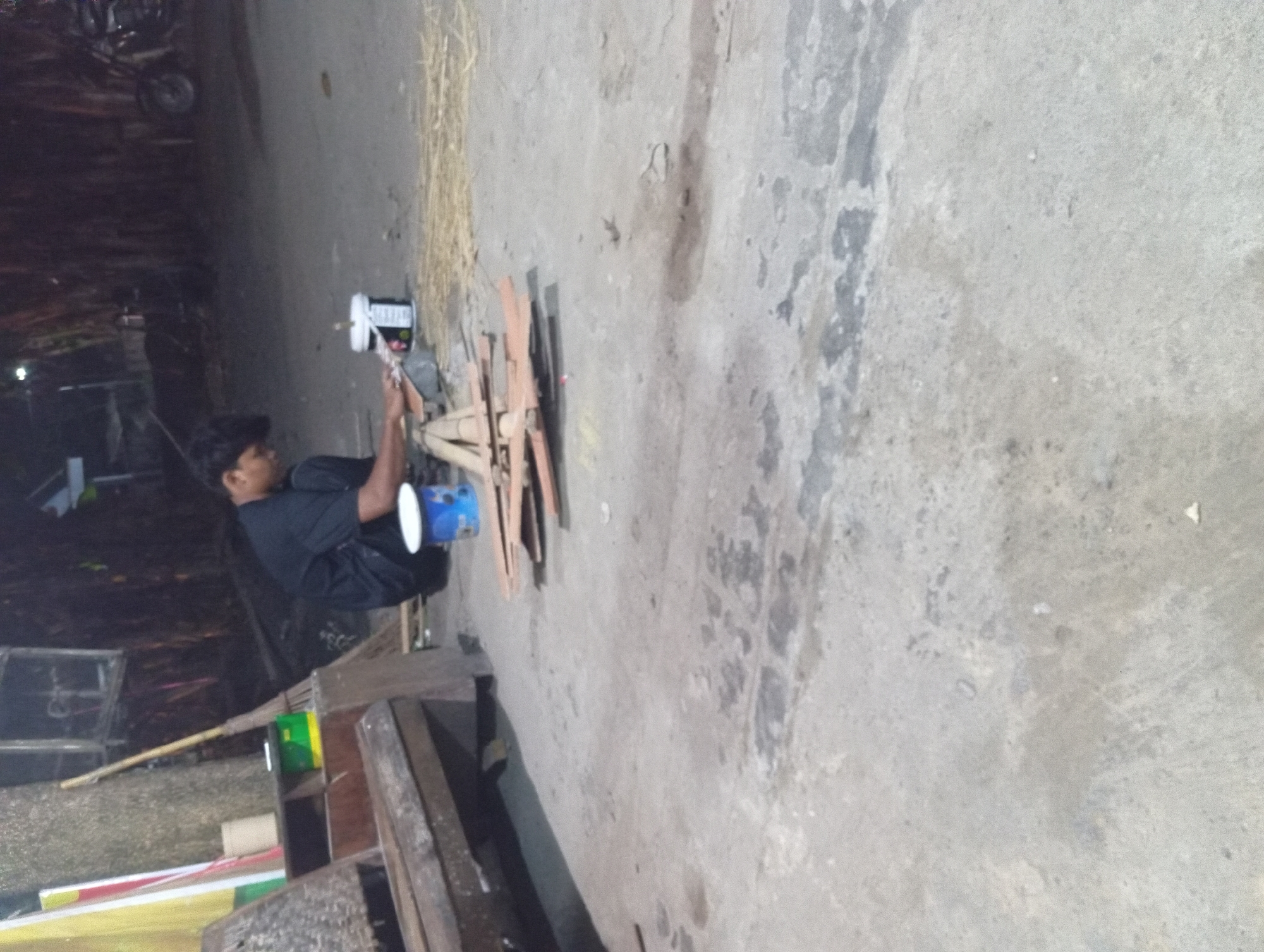 The image depicts an outdoor scene in Sidokarto, Indonesia, at night. A man, dressed in a black shirt and pants, is seated on the ground, working on several flat, wooden objects. These objects appear to be unfinished or in the process of being assembled, with some stacked on top of each other. In front of the man, there is a blue and white bucket, possibly containing paint or adhesive. To the right of the bucket, there is a small pile of dried straw or hay. The ground is paved with concrete, showing some patches of discoloration and dampness. In the background, a wooden structure with a broom leaning against it is visible, along with a motorcycle parked to the right. The overall setting suggests a workshop or a storage area.