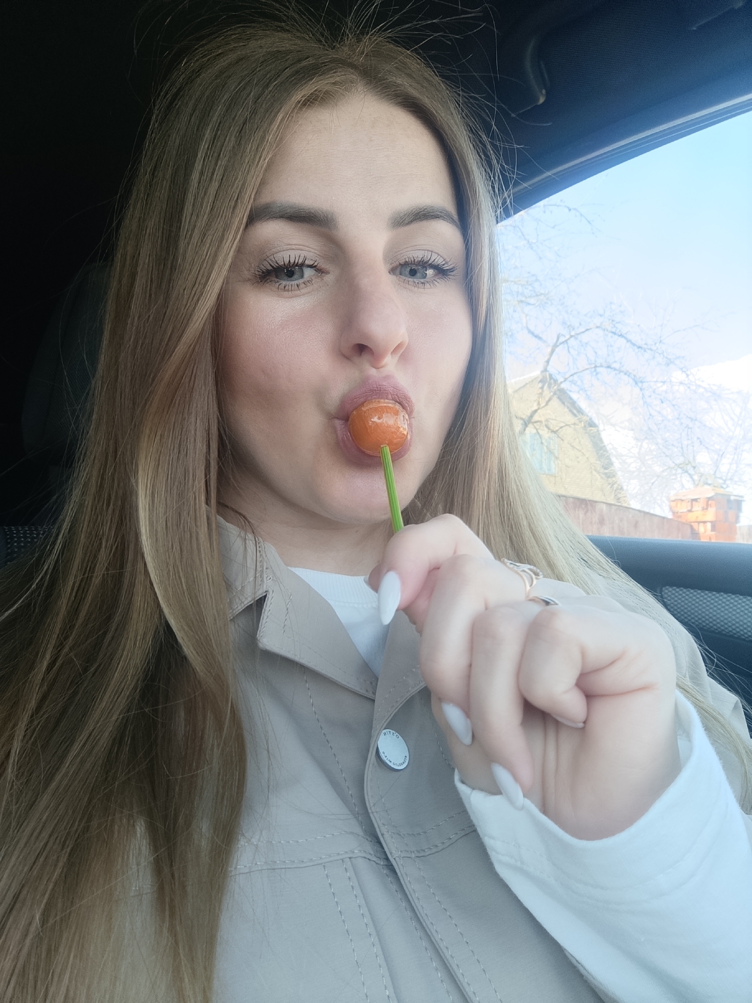 A woman with long, light brown hair is sitting in a car, holding an orange lollipop in her mouth. She is wearing a beige jacket over a white shirt. Her fingernails are painted white. Outside the car window, there is a building and a tree. The location is the city of Unknown, Unknown.