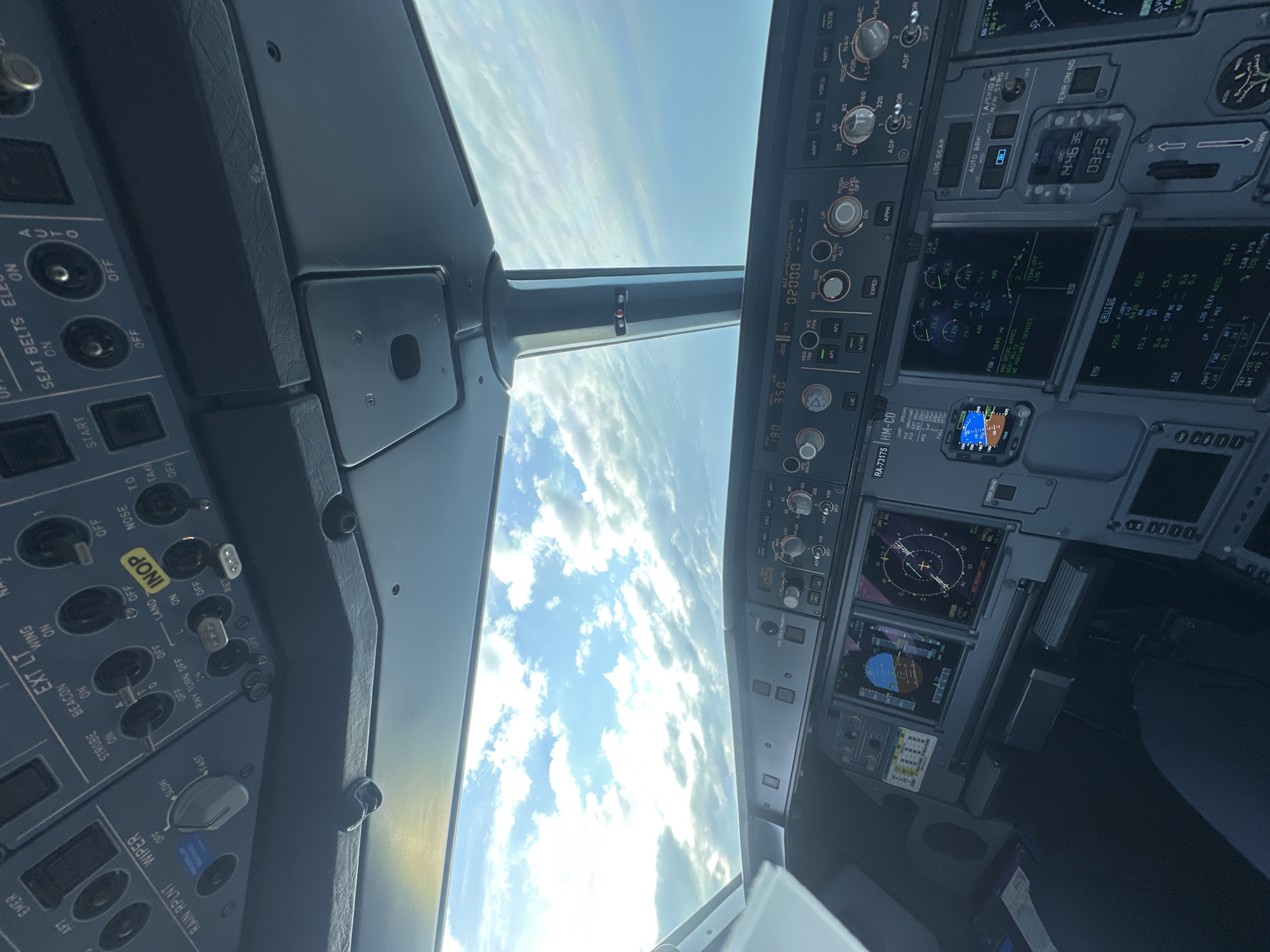 The image shows the interior of an aircraft cockpit in flight. The view through the windscreen reveals a sky with scattered clouds. The control panel displays a variety of instruments and gauges, indicating flight parameters. Switches related to lights, wipers, and other systems are visible. Some text indicates "RAIN RPLNT", "WIPER", "STROBE", "BEACON", "EXT LT", "WING", "RWY TURN OFF", "LAND", "NOSE", "TAXI", "START", "SEAT BELTS ELE", "AUTO", "VOR2", "HM-CD", "HDG", "V/S", "LOC", "A/THR", "AP2", "METRIC", "ALT", "APPR", "CRUISE", "ENG", "F.USED", "ARPT", "VORD", "NAV", "ARC", "ADF", "LOG GEAR", "AUTO BRK", "A/S", "SKID", "STRG", "TERR ON ND", "AIR". The aircraft tail number is RA-73175. The location and city of Sochi, Russia cannot be ascertained from the image.