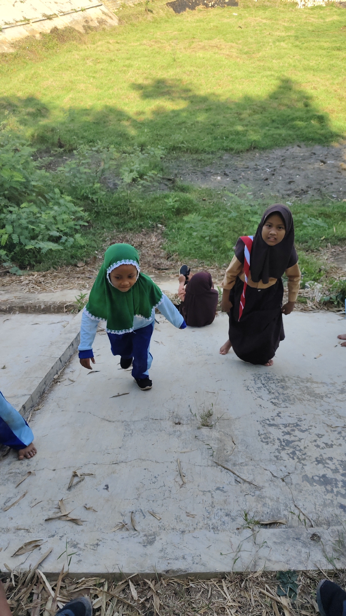 Four young girls, dressed in Islamic school uniforms, are on a concrete path outdoors. The girl in the foreground on the left wears a green hijab and a light blue shirt with dark blue pants. She is walking forward with her head turned slightly to her left. Another girl, to her right and slightly behind, wears a dark brown hijab and dress with a red and white sash. She is facing forward and looking at the camera. Behind them, two other girls are visible: one is seated on the ground, wearing a dark brown hijab and the other is partially visible at the far right of the frame. The concrete path is cracked and uneven, with dried leaves and grass scattered on it. In the background, there is a grassy slope with some vegetation. The scene appears to be in a somewhat unkempt outdoor area, possibly a schoolyard or recreational space.