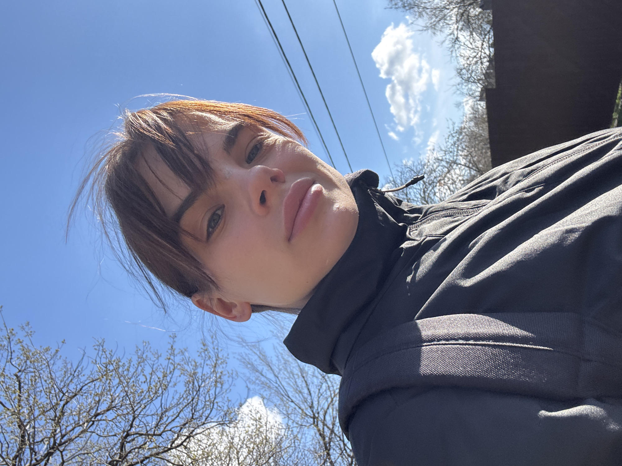 A low-angle, tilted selfie shot shows a fair-skinned woman with shoulder-length brown hair and bangs, wearing a dark-colored jacket with a high collar and a backpack, against a background of a bright blue sky with wispy clouds and the bare branches of trees. Overhead power lines are visible in the upper portion of the image. The photo's composition suggests the subject is outdoors, possibly in a park or wooded area. There's no way to confirm or deny that this photo was taken in Rostov-on-Don, Russia.