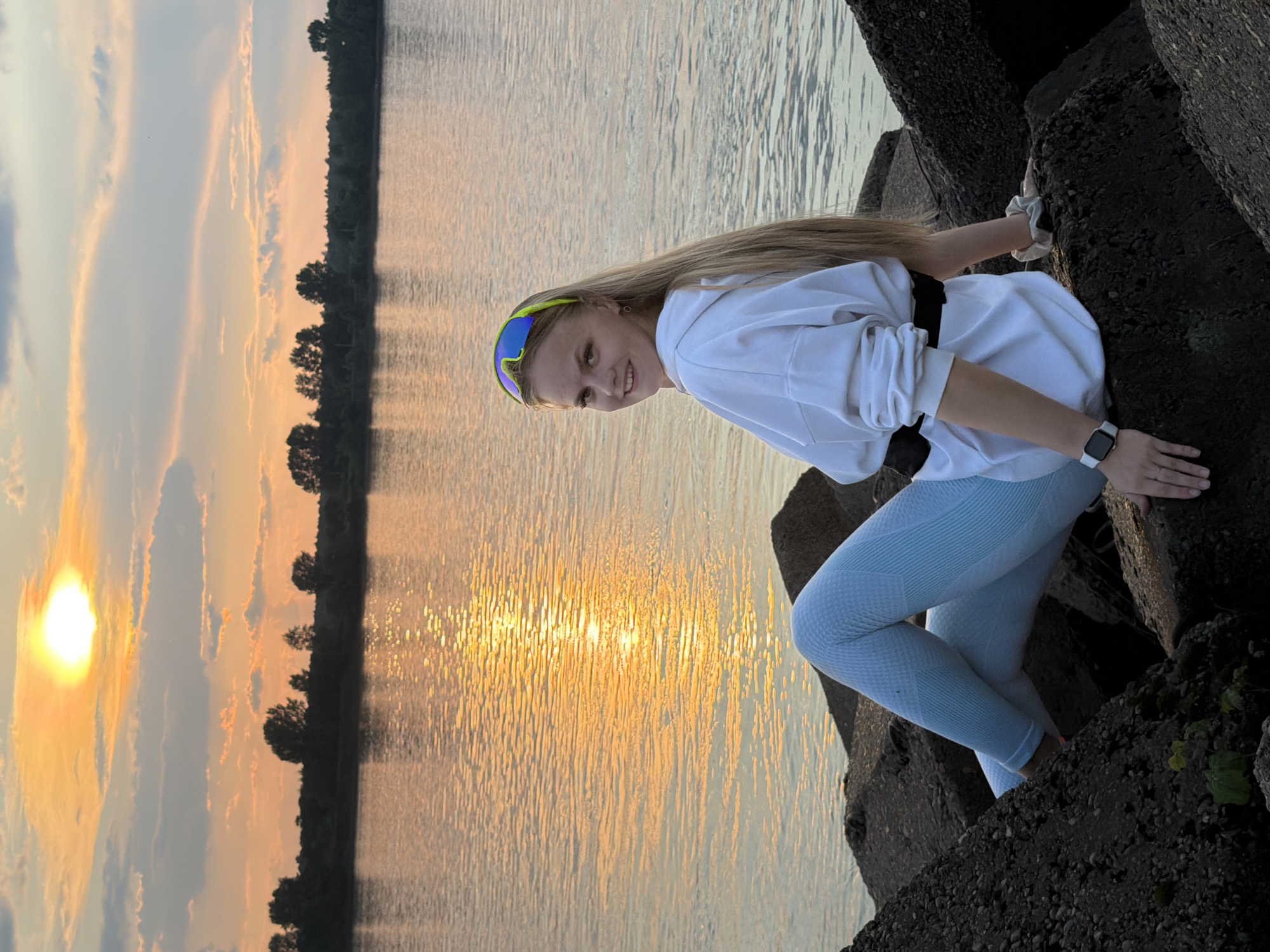 A young woman with blonde hair and sunglasses is sitting on large rocks by a body of water at sunset. The sun is low in the sky, casting a warm orange glow on the clouds and the water. The woman is wearing a white hoodie and light blue leggings. She is looking over her shoulder at the camera with a smile. The background shows a treeline across the water. This scene is likely at the Kama River embankment in Chaikovsky, Russia, a popular spot for recreation and enjoying the natural scenery.