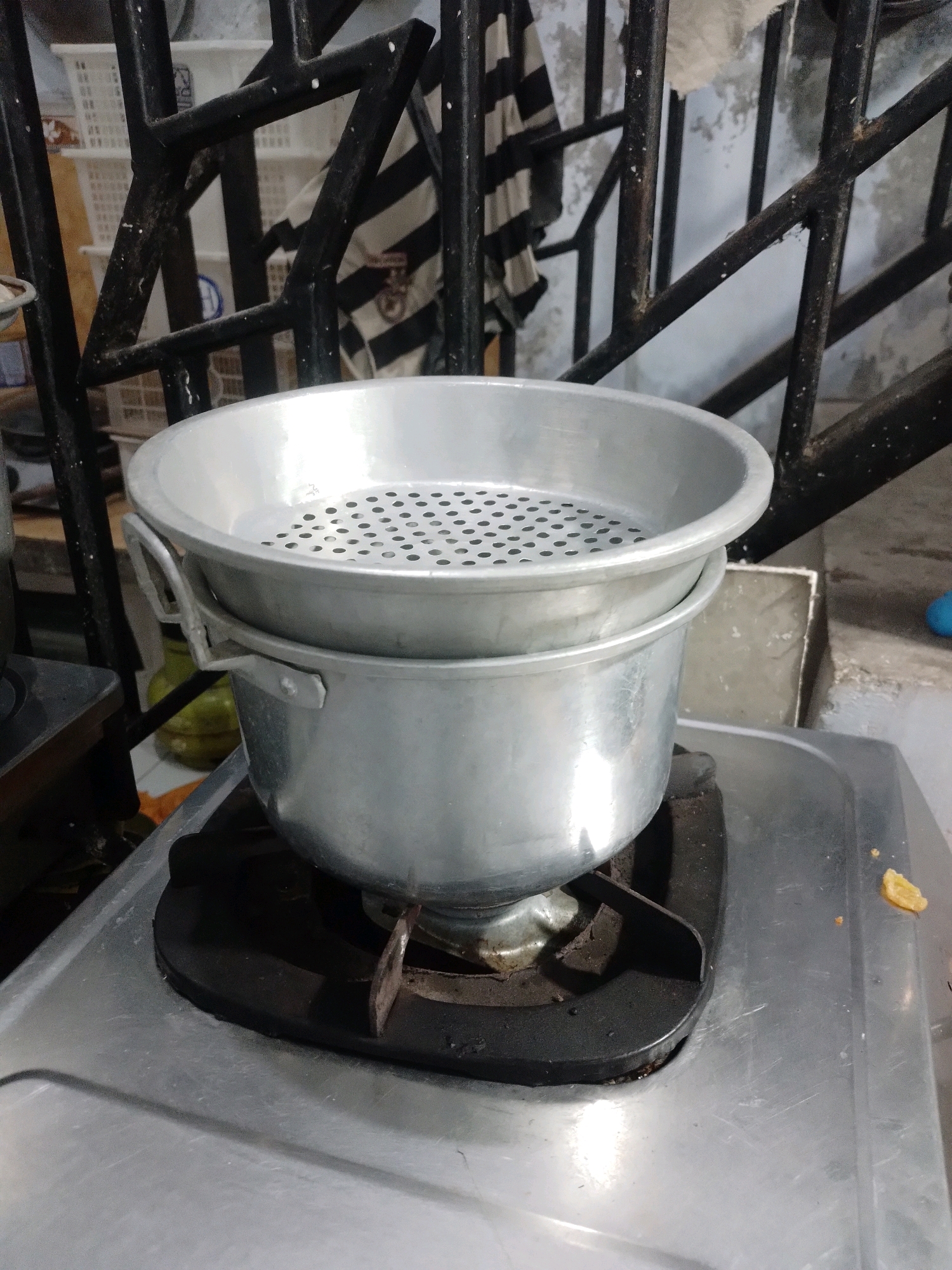 The image shows a two-tiered aluminum steamer pot placed on a gas stove in Pelem, Indonesia. The steamer pot is silver and has a perforated tray on top. The gas stove is also silver and appears to be in a kitchen setting. In the background, there is a black metal railing, a striped cloth hanging, and some white plastic baskets. The overall scene suggests a domestic environment where food is being prepared.