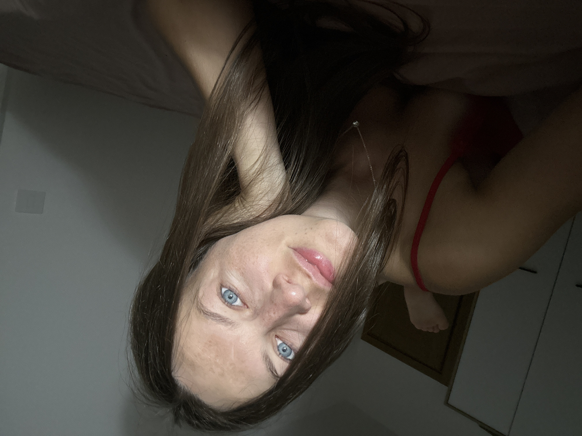 A young woman with long brown hair and blue eyes is lying on a bed, propped up on her elbow. She is wearing a red bra and a silver necklace. The room is dimly lit, with a wardrobe and a doorway visible in the background. The image appears to have been taken from a close-up perspective, focusing on the woman's face and upper body. The location is stated to be Donji Pobori, Montenegro.