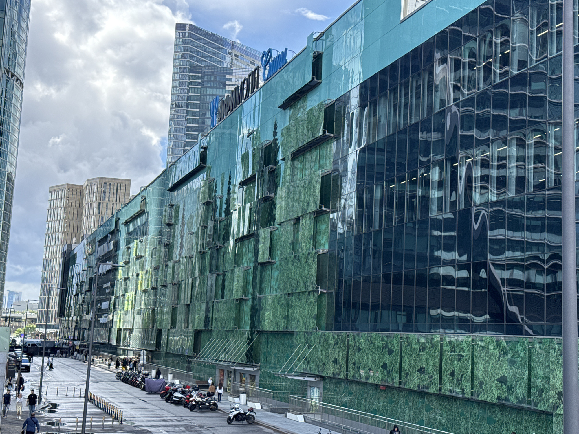 This is a wide, eye-level shot of a modern building in Moscow, Russia. The building is clad in green, reflective panels that show a distorted reflection of trees and sky. The building has several stories with rows of windows. Above the green panels, the building is covered in dark blue, reflective panels that show distorted reflections of other buildings and the sky. A blue sign with white lettering is visible at the top of the building. In the foreground, a street is lined with streetlights and a number of motorcycles are parked. People are walking on the street and sidewalks. In the background, other tall modern buildings are visible, along with a cloudy sky.
