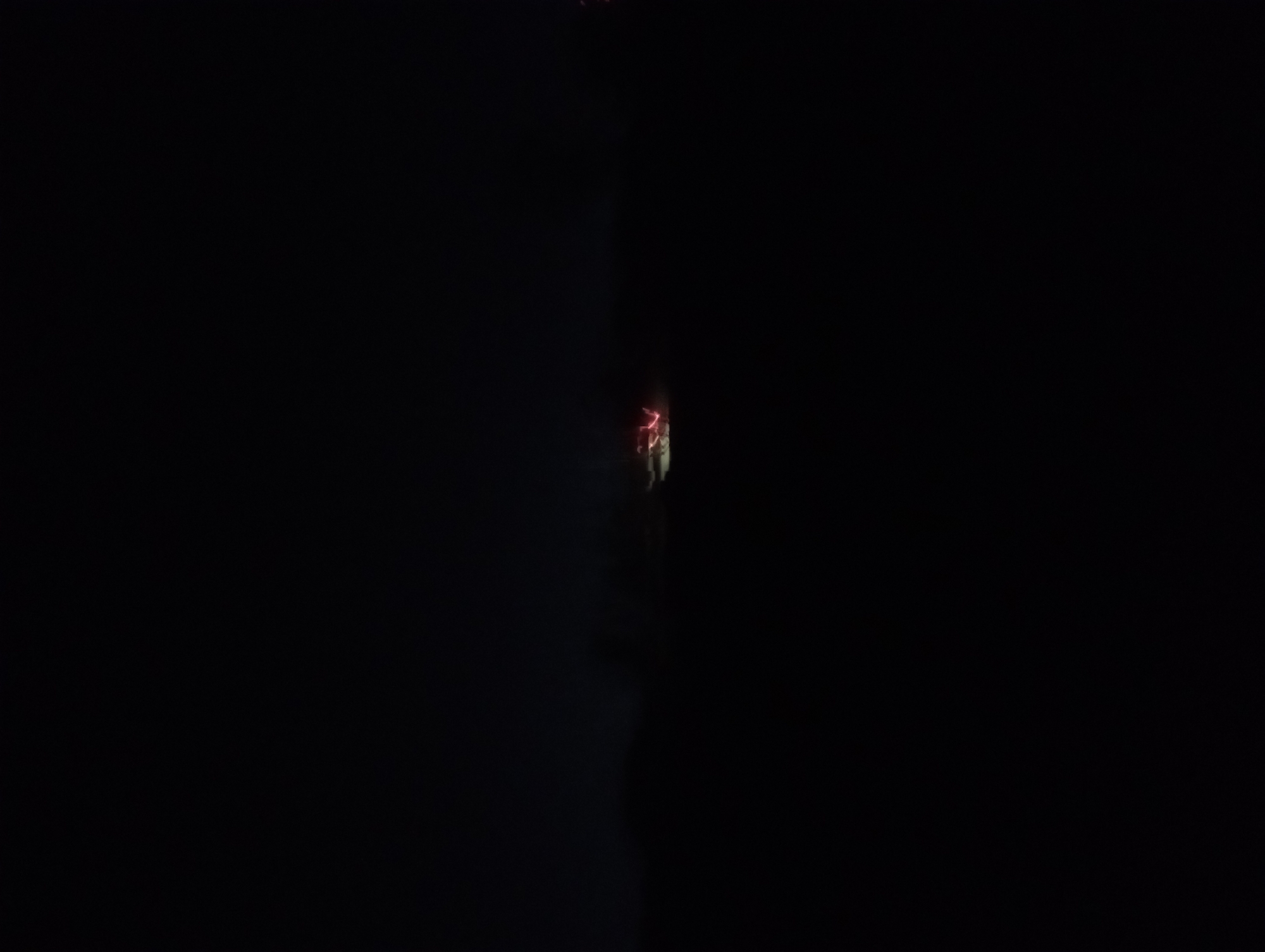 The image depicts a dark, nighttime scene with a prominent silhouette of a low-lying horizon, possibly indicating a natural landscape or the outskirts of a city. In the middle ground, a distinct cluster of bright red and pink lights is visible, suggesting the presence of artificial illumination. These lights appear somewhat abstract and blurred, possibly due to movement or atmospheric conditions. The overall impression is of a distant light source or activity occurring in the darkness, set against a black, featureless sky. There are no clear indications of specific buildings, vehicles, or individuals in the image. Without additional context, it is impossible to definitively determine the location within Reifild-Ist, Nigeria.