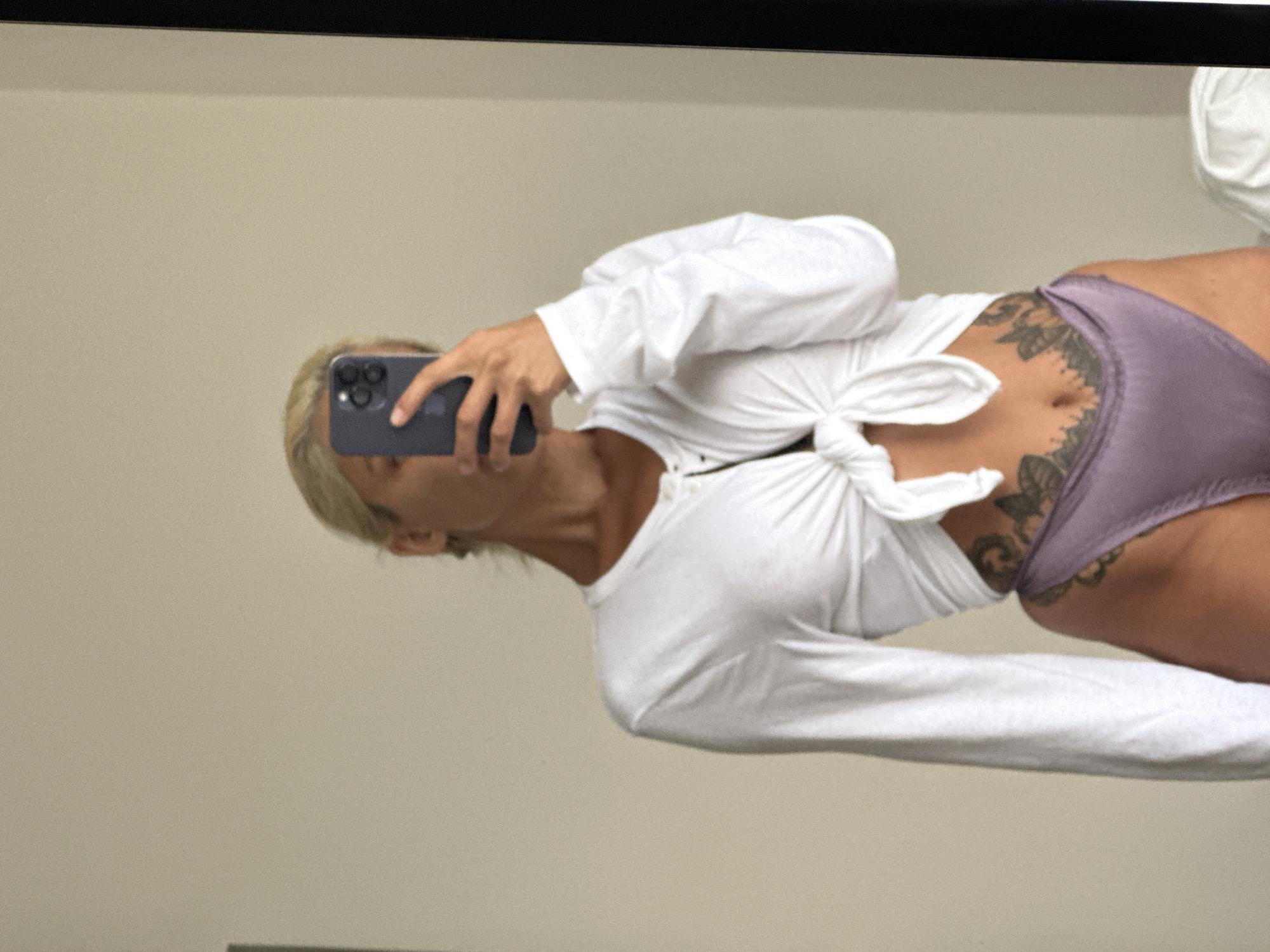 A woman with blonde hair is taking a mirror selfie in what appears to be a bedroom. She is wearing a white long-sleeved shirt tied at the waist and purple underwear. Her abdomen is visible, displaying intricate tattoos. The background is a plain wall, and a black framed mirror reflects the scene. There is a glimpse of a bed with white sheets in the lower right corner of the reflection. The lighting suggests it is daytime, possibly indoors with natural light. This scene is likely in a private residence or hotel room in Kuta Selatan, Indonesia.